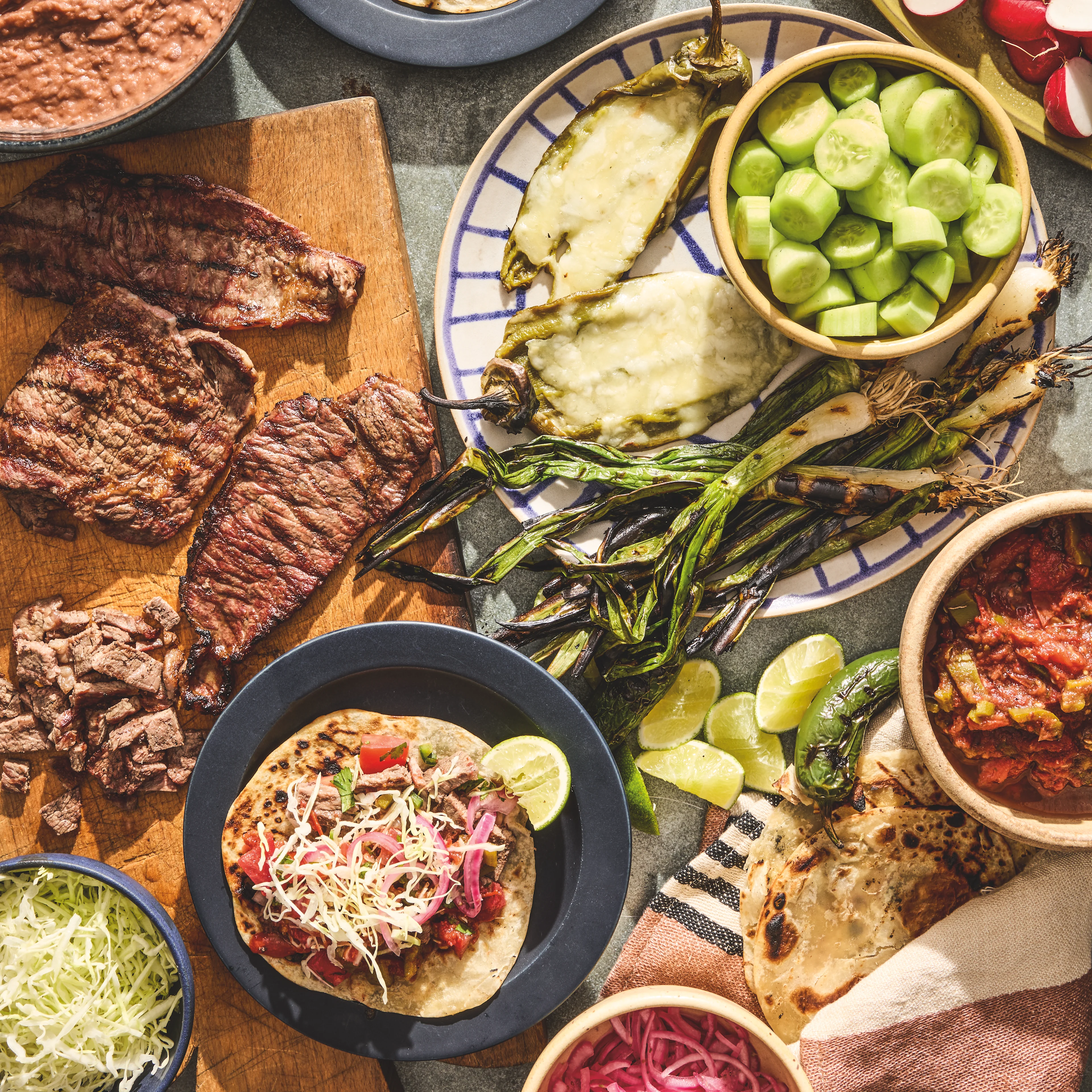 A spread of Mexican food including grilled steak, tacos, roasted peppers, cucumber, pickled onions, salsa, and refried beans.