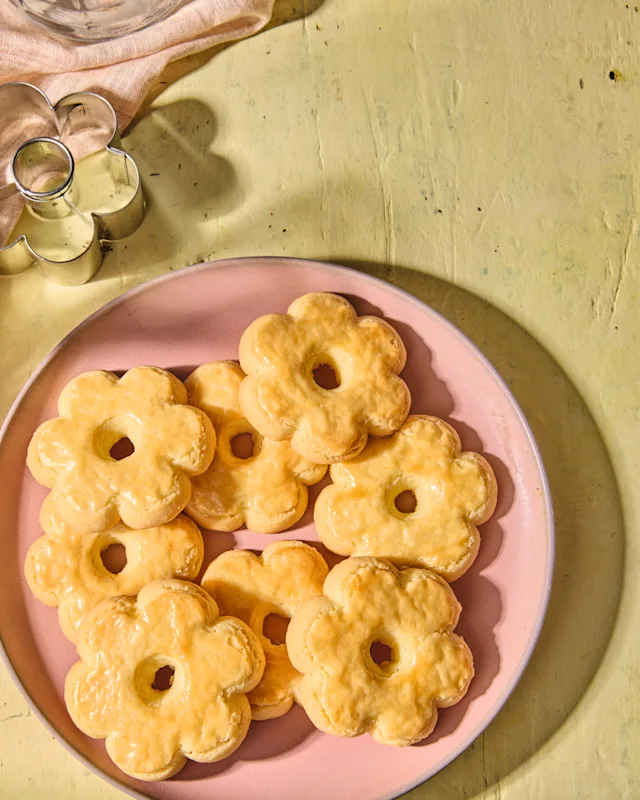 Golden flower-shaped donuts piled on a pink plate, set on a yellow surface with cookie cutters nearby.