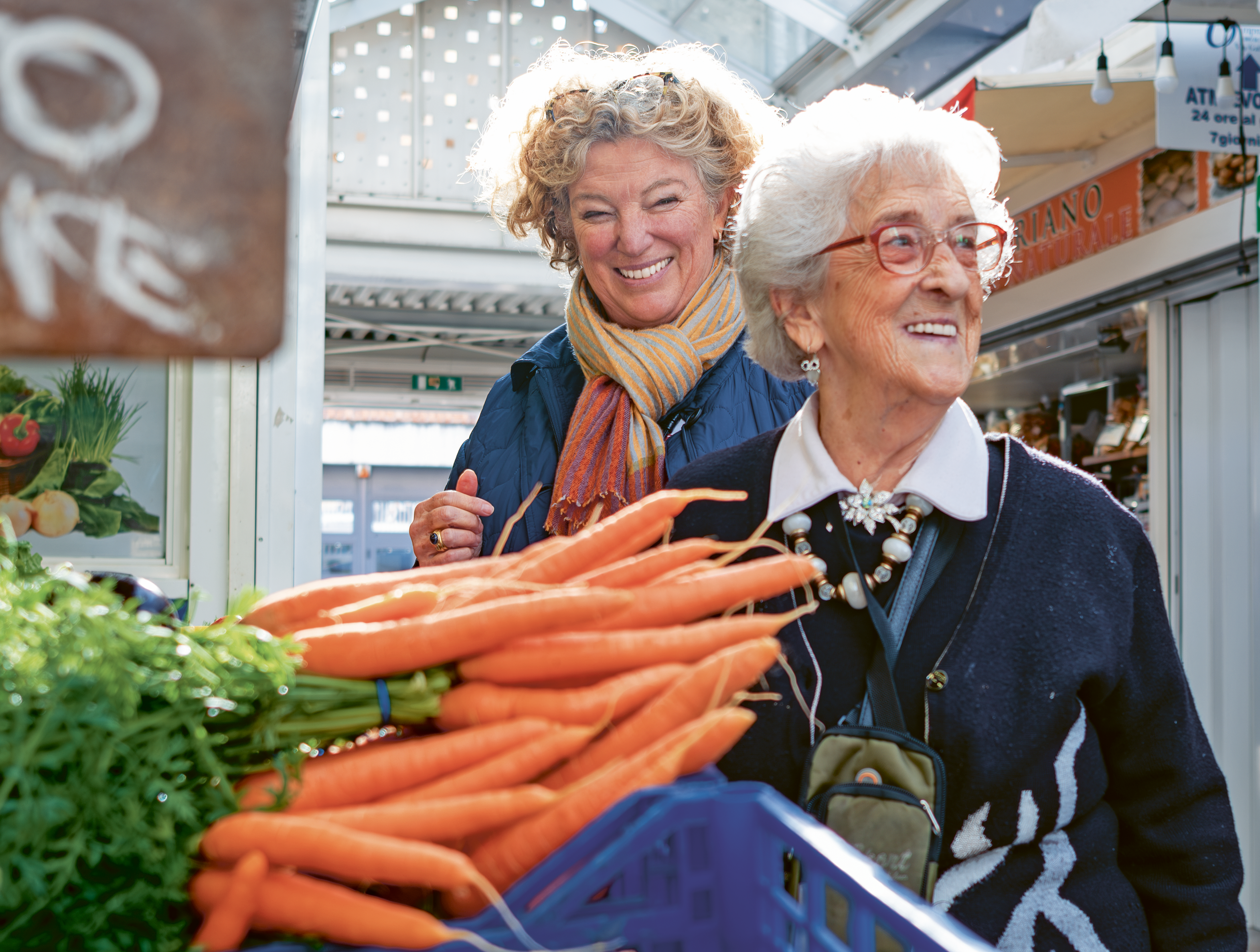 Two smiling women browse fresh carrots at an outdoor market, one elderly with red glasses, the other middle-aged with a colorful scarf.