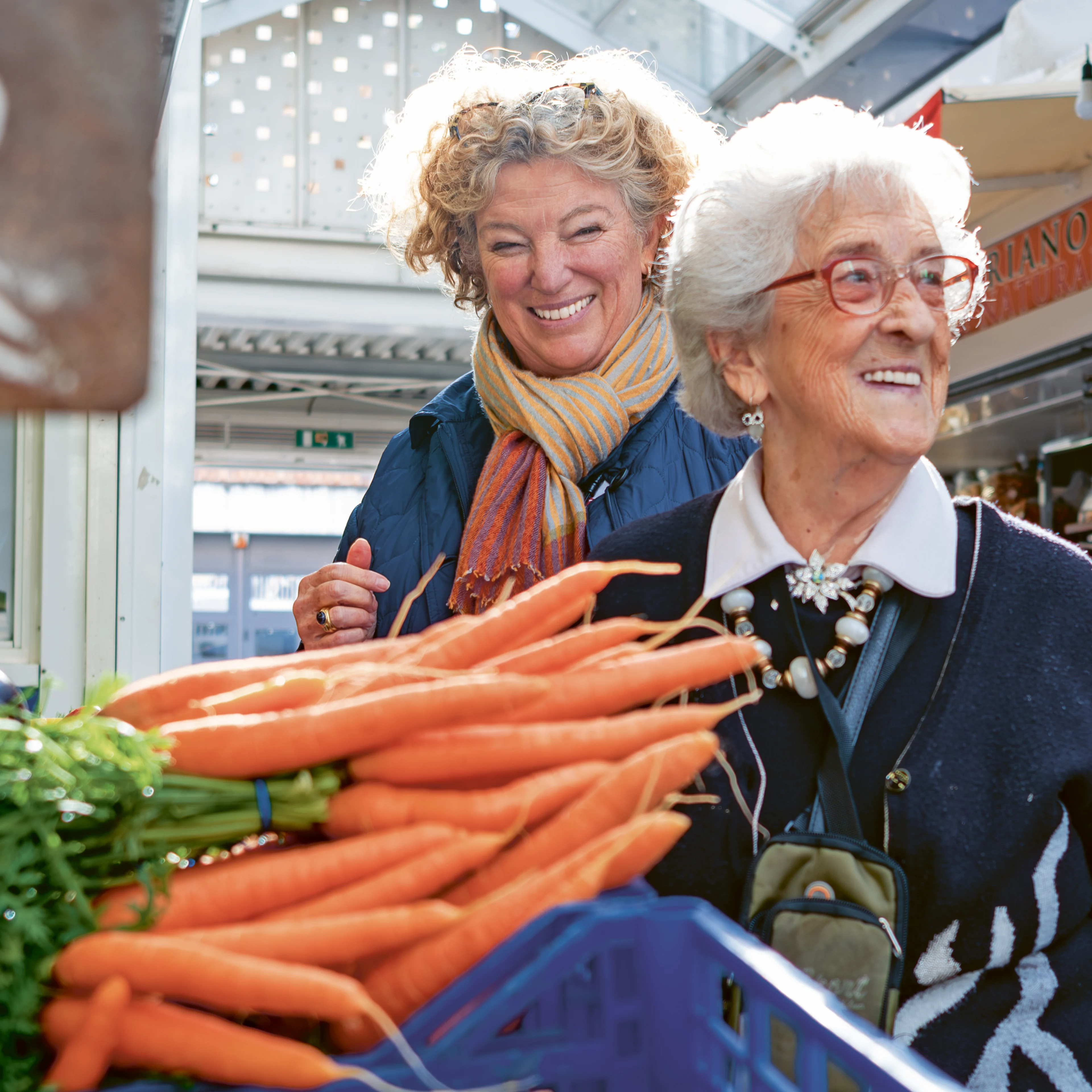 Two smiling women browse fresh carrots at an outdoor market, one elderly with red glasses, the other middle-aged with a colorful scarf.