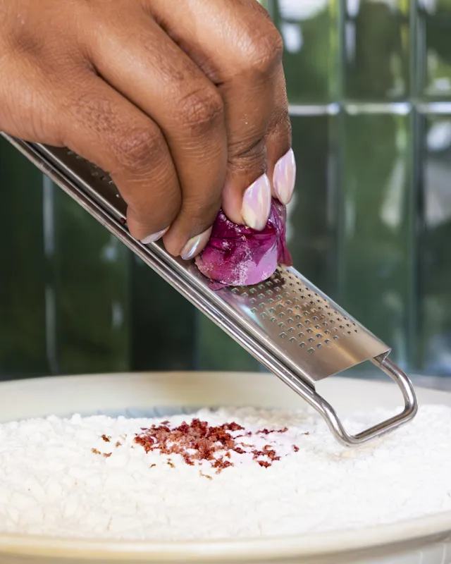 A hand grating a red onion over a white plate of flour using a stainless steel grater.