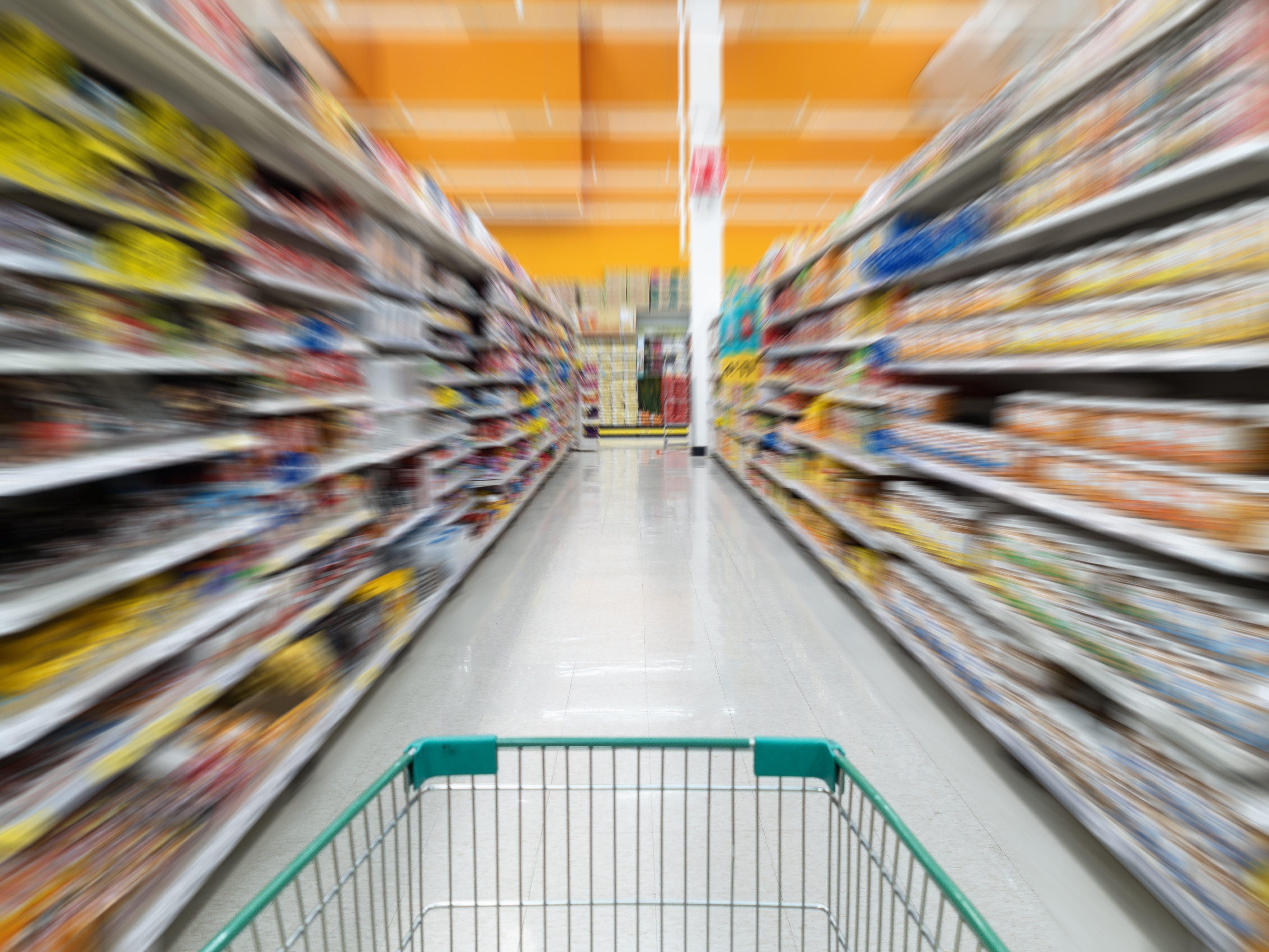 Blurred grocery store aisle view from shopping cart perspective, showing motion through shelves under orange ceiling