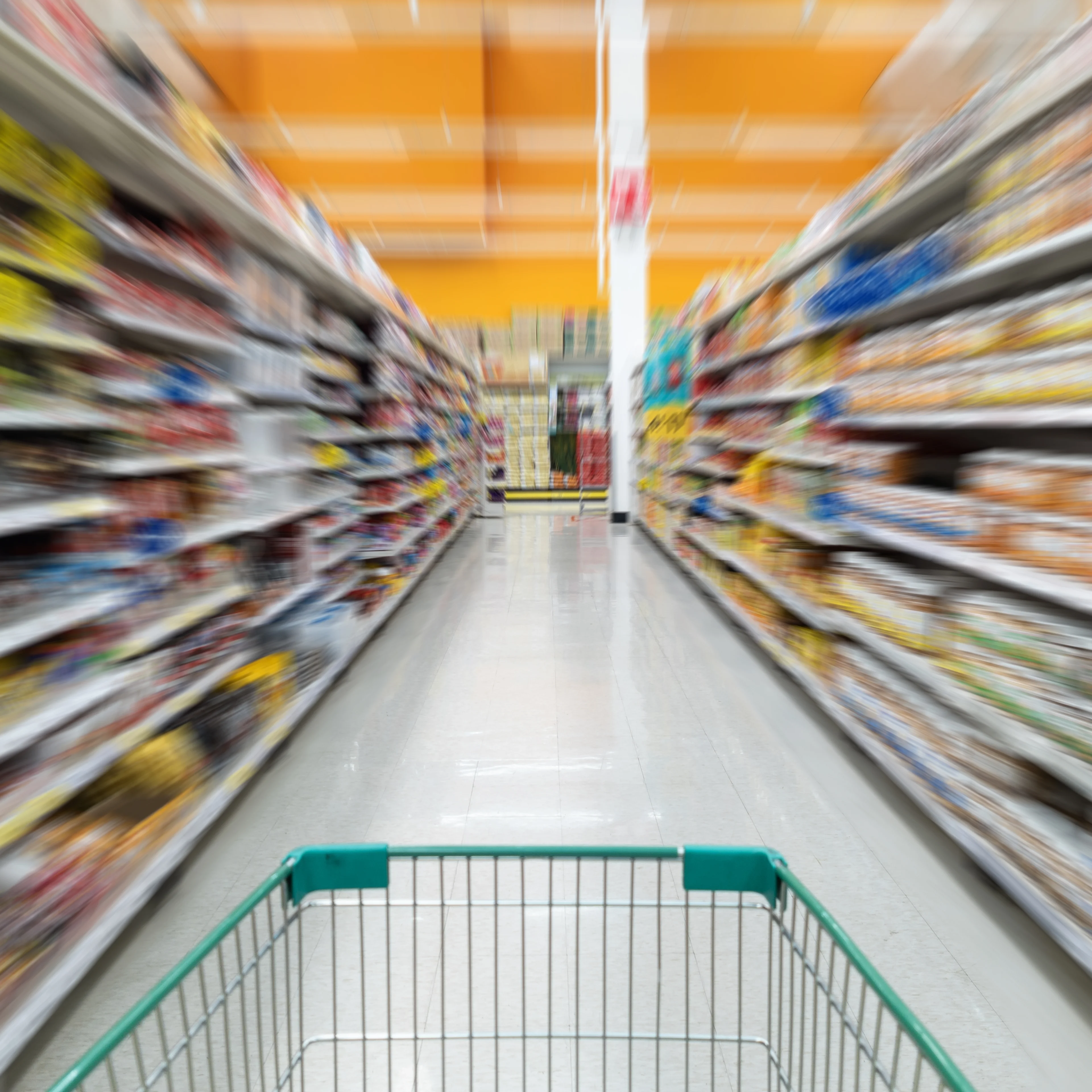 Blurred grocery store aisle view from shopping cart perspective, showing motion through shelves under orange ceiling