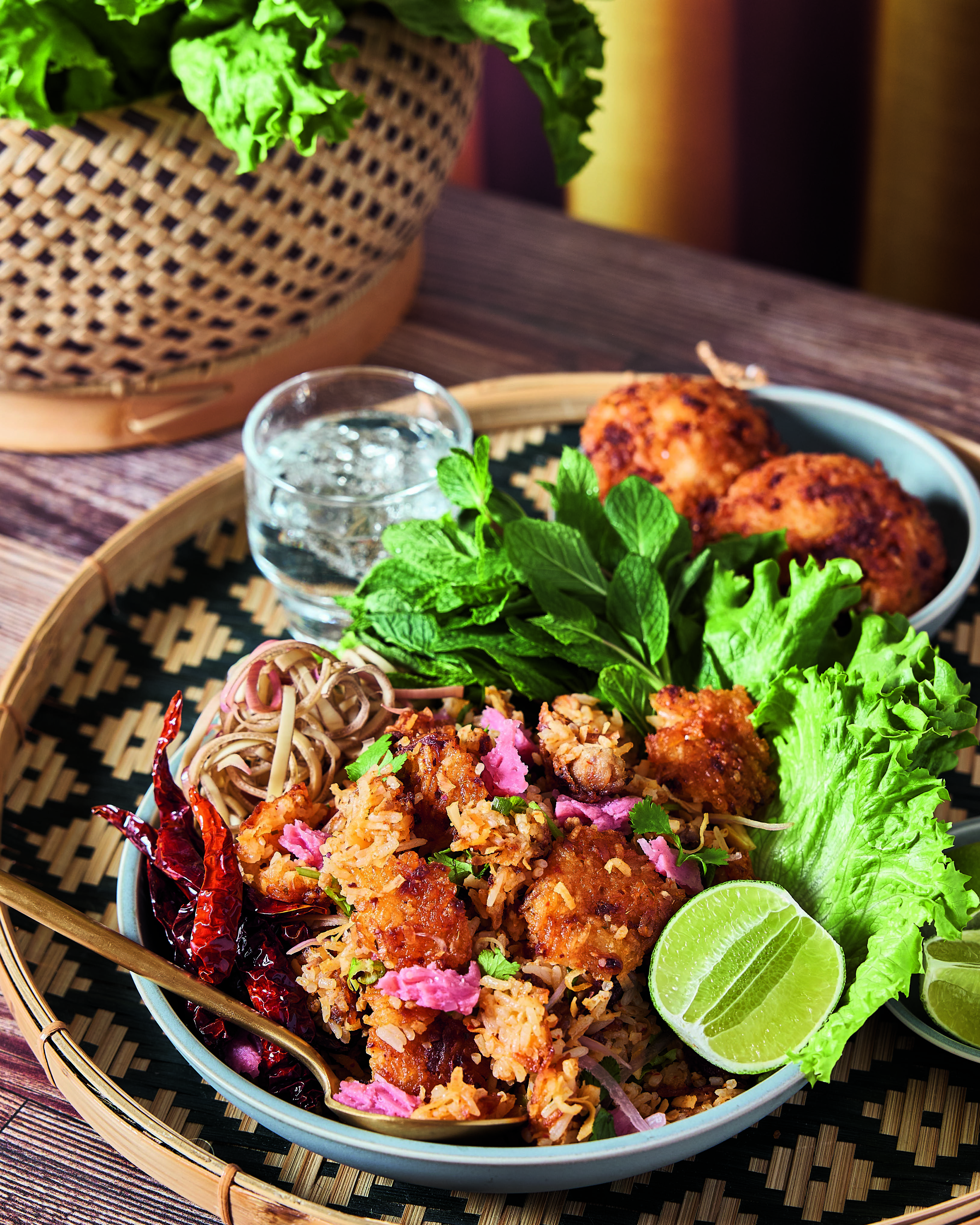 A plate of fried rice with crispy chicken, dried chilies, lime, fresh herbs, lettuce, and noodles on a woven tray.