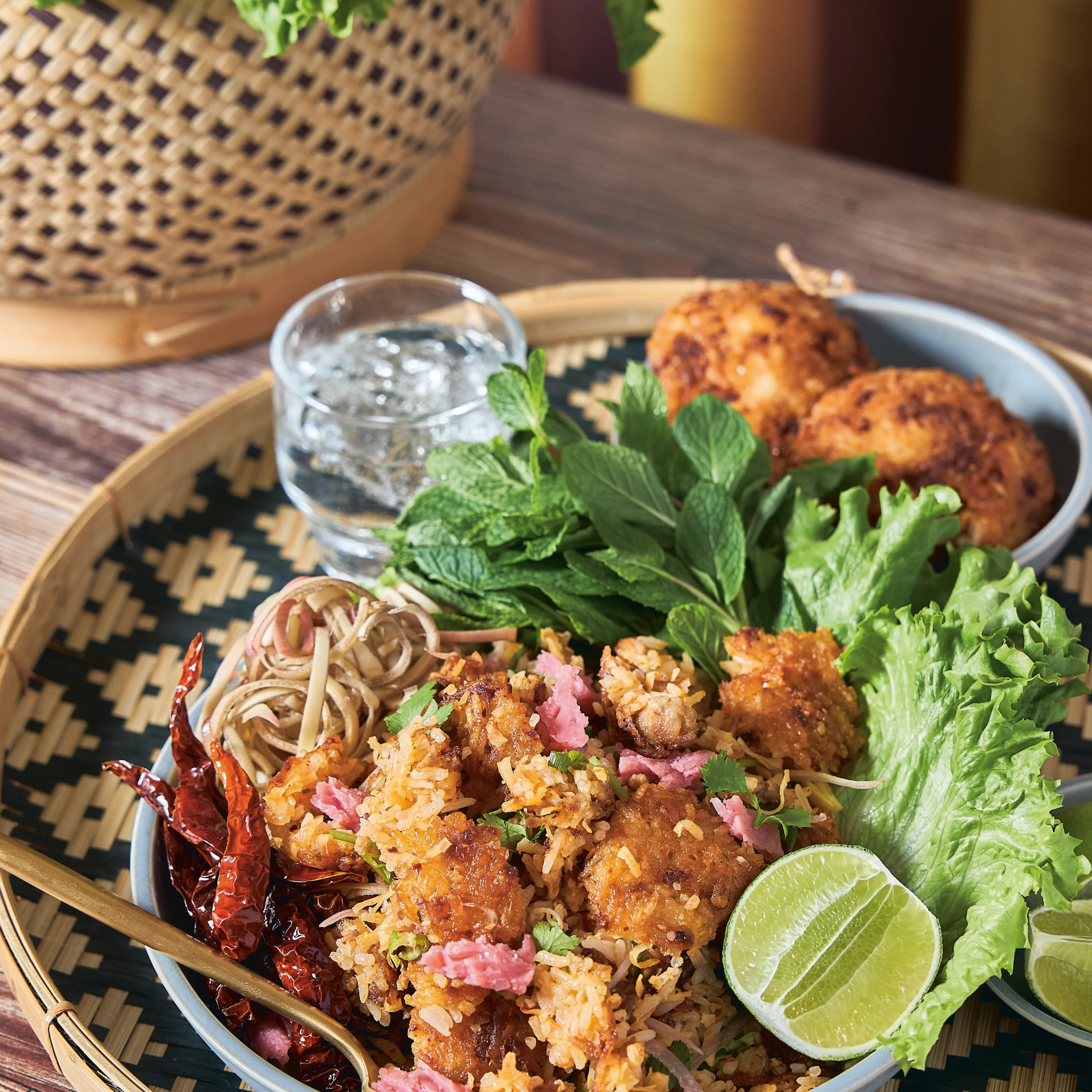 A plate of fried rice with crispy chicken, dried chilies, lime, fresh herbs, lettuce, and noodles on a woven tray.