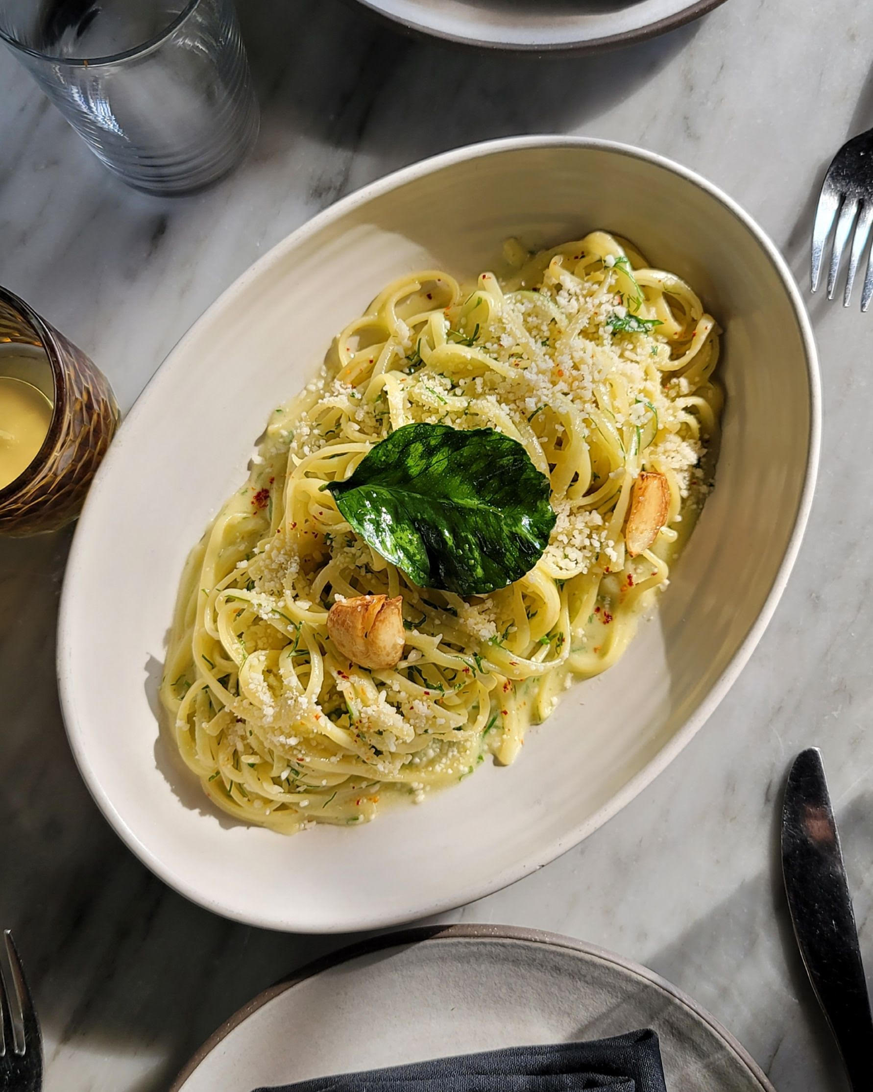 Pasta dish in white bowl with parmesan cheese and basil leaf garnish on marble table with water glass and utensils.