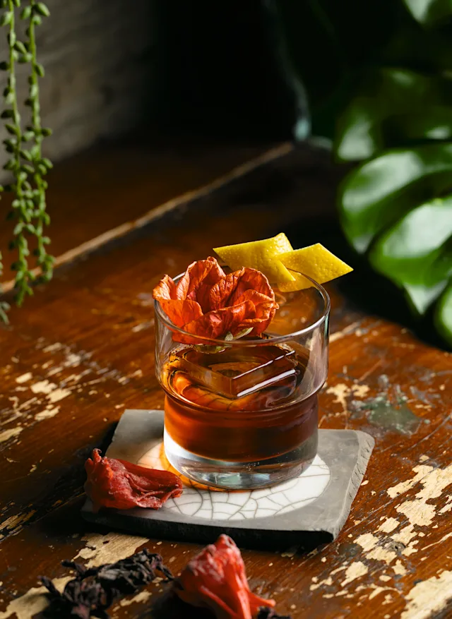 A amber cocktail in a rocks glass garnished with a dried red flower and lemon peel, resting on a ceramic coaster on a wooden surface.