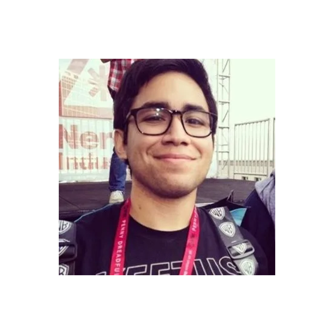 A young person with glasses and a red lanyard smiling at an outdoor event with stage equipment visible in background.