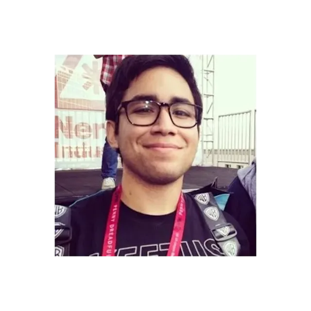 A young person with glasses and a red lanyard smiling at an outdoor event with stage equipment visible in background.