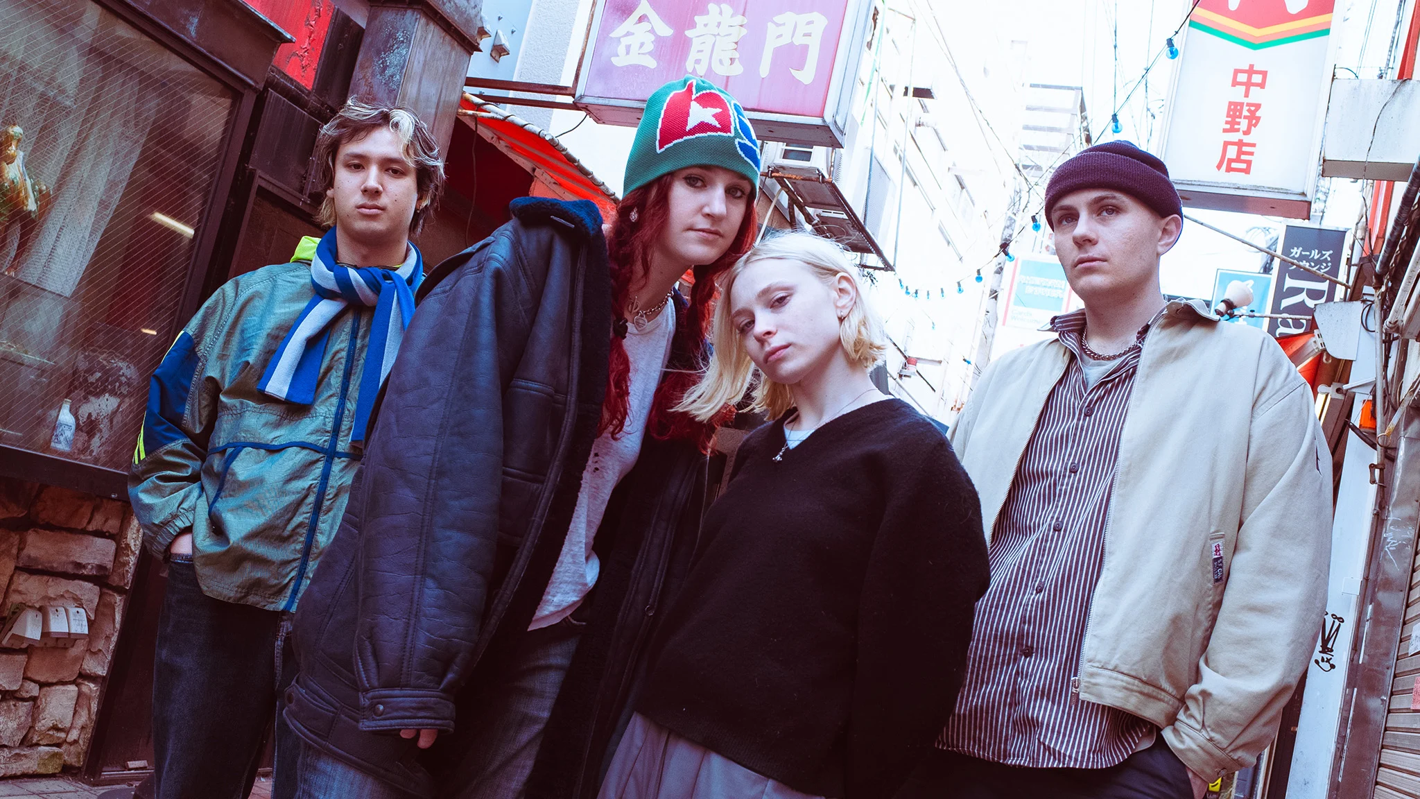 Four young people pose in a narrow Japanese alley lined with colorful signs, dressed in casual streetwear.