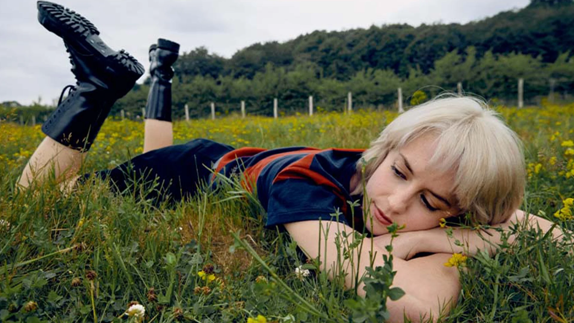 Avalon Emerson lies in a green field of wildflowers, wearing a navy top and black boots, resting her head on her arms.
