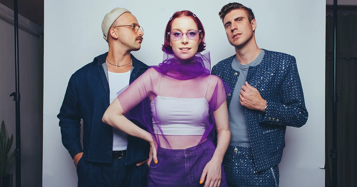Three stylish musicians known as Moonchild posing together, woman in purple sheer top with red hair between two men in fashionable attire.