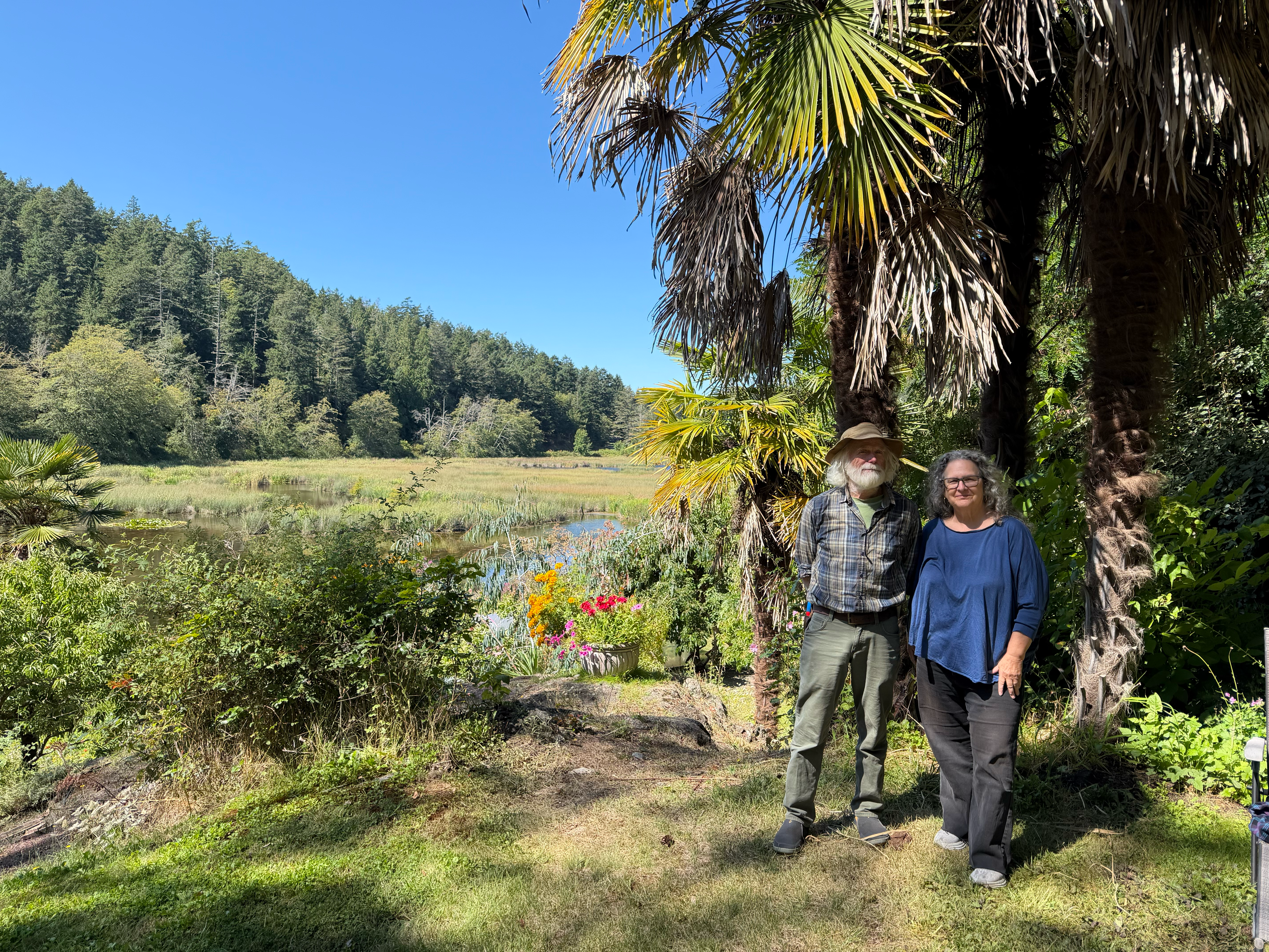 Two people stand near palm trees in a lush garden with colorful flowers, a wetland, and forested hills under a blue sky.