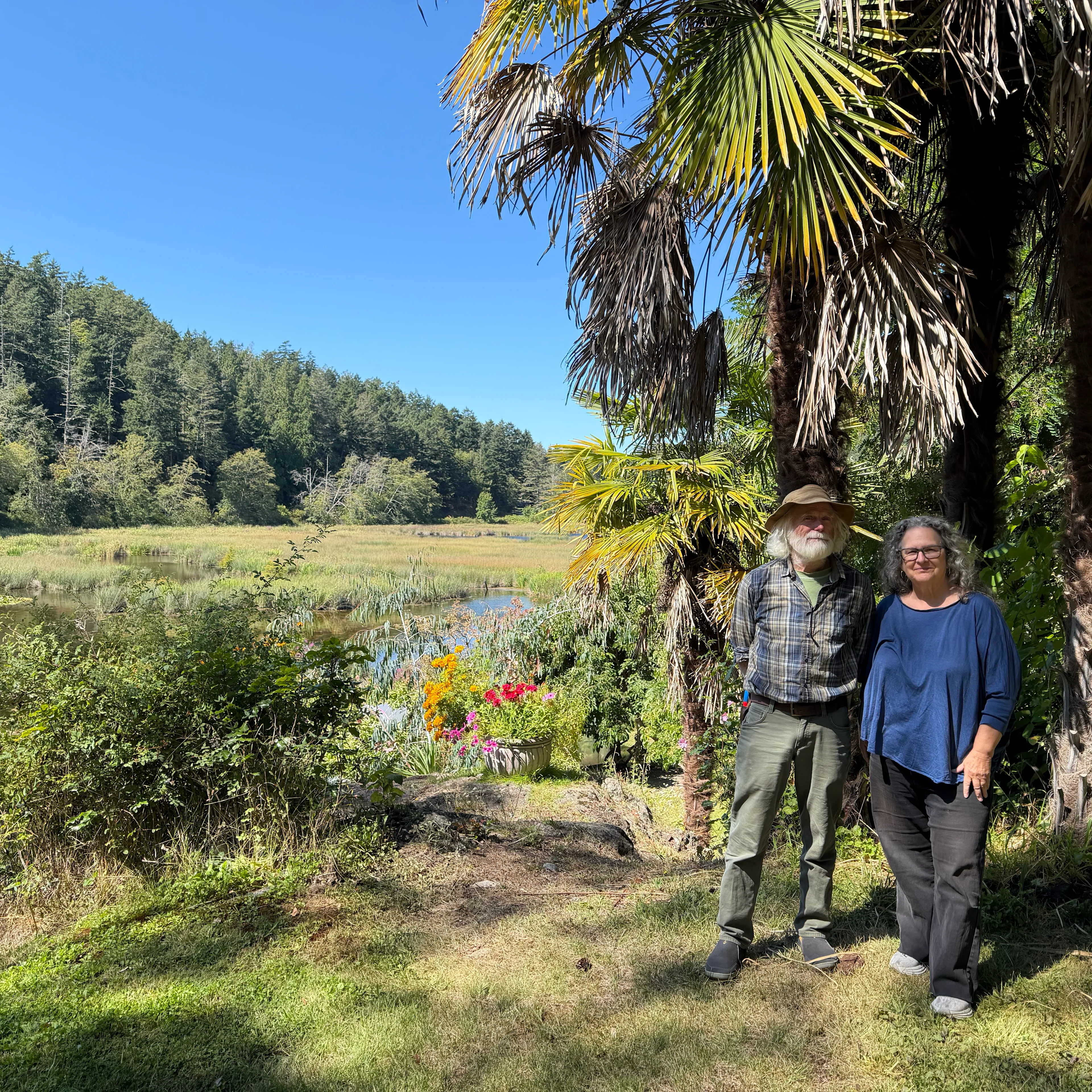 Two people stand near palm trees in a lush garden with colorful flowers, a wetland, and forested hills under a blue sky.