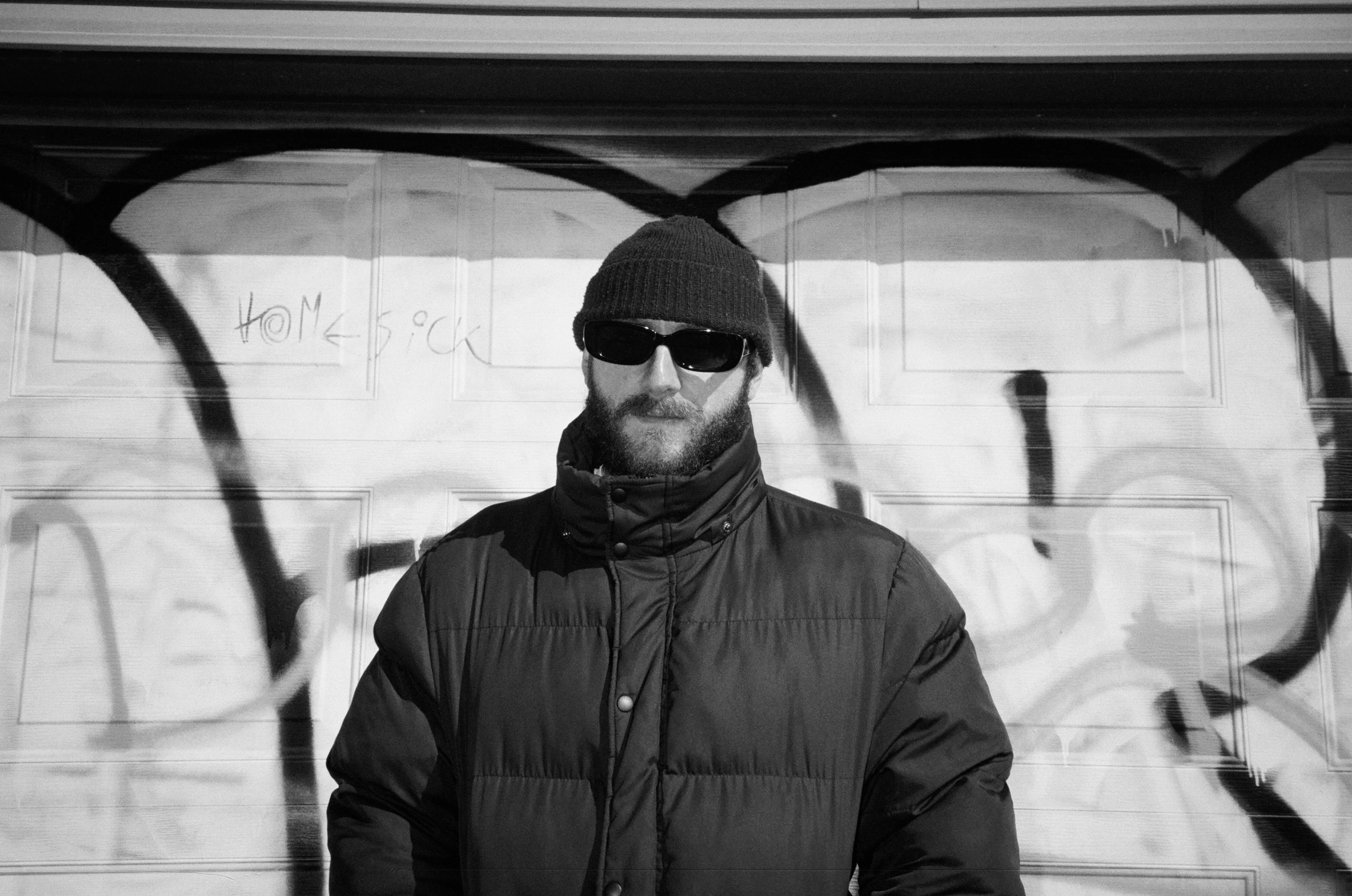 A bearded man in a beanie and sunglasses wearing a puffer jacket, standing before a graffiti-covered wall in a black and white photo.