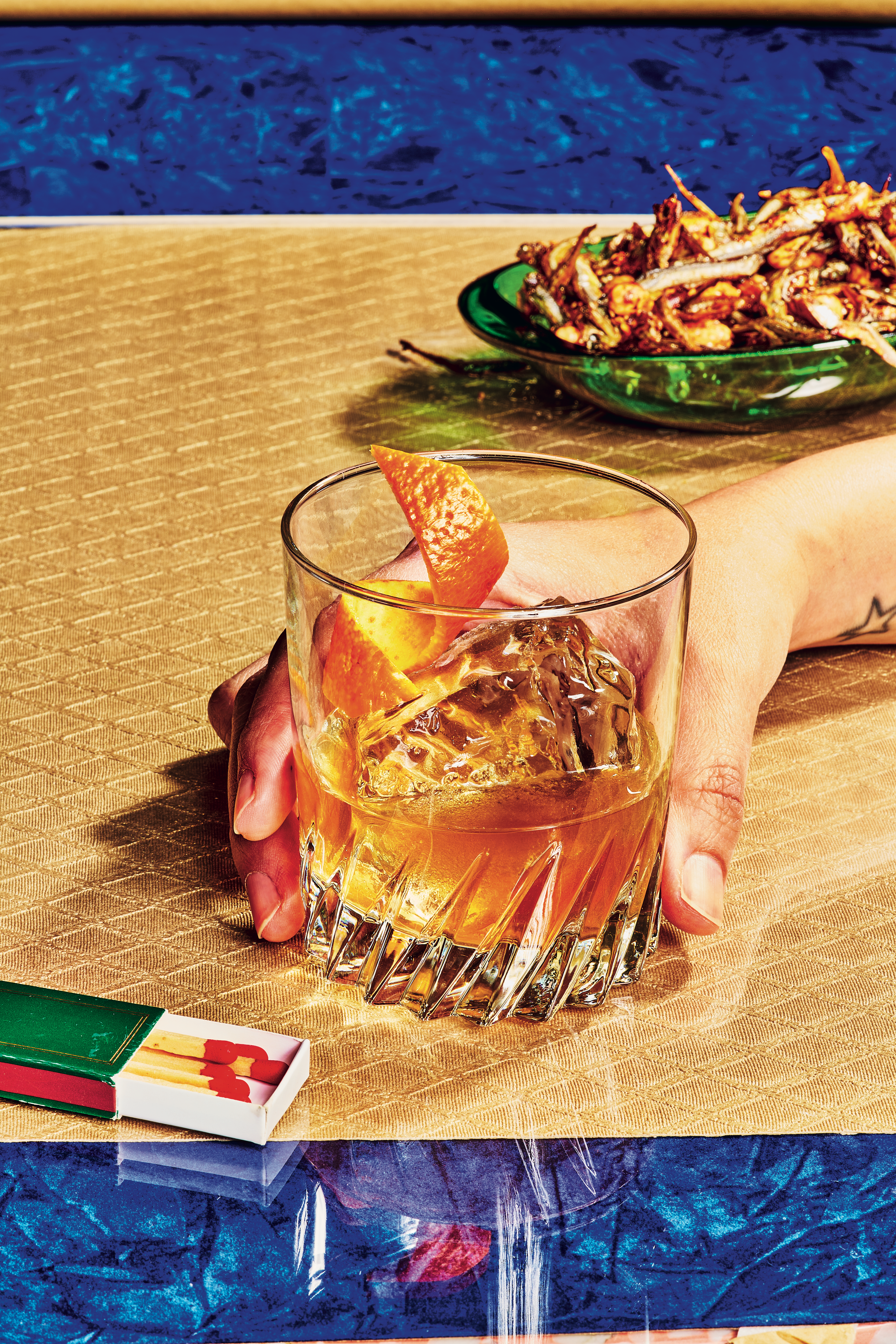 A tattooed hand holds an amber cocktail with orange peel on a gold surface, beside matchbooks and a green plate of fried food.