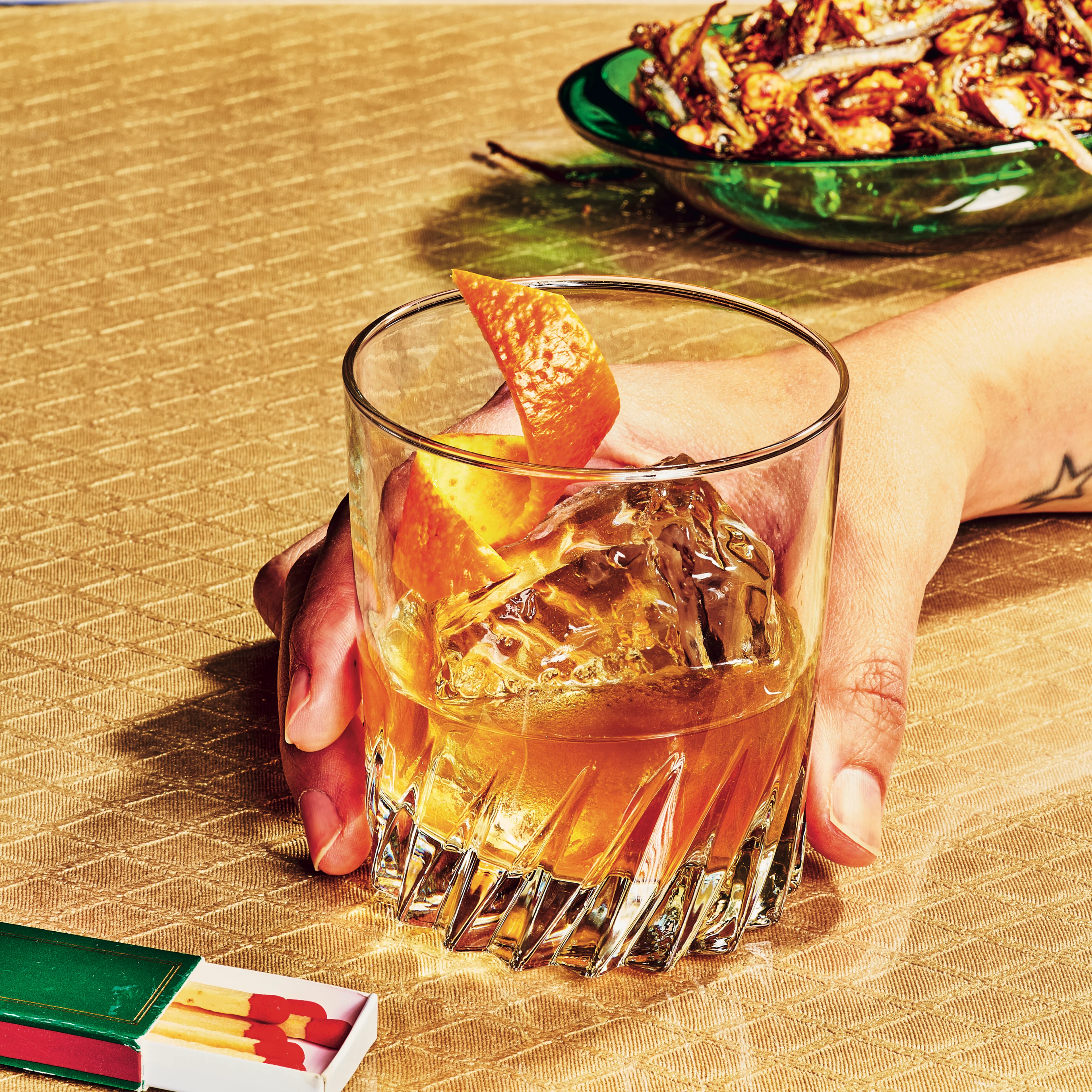 A tattooed hand holds an amber cocktail with orange peel on a gold surface, beside matchbooks and a green plate of fried food.