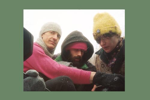 Three people in winter clothing huddled together outdoors, wearing knit hats and hooded jackets in cold weather.