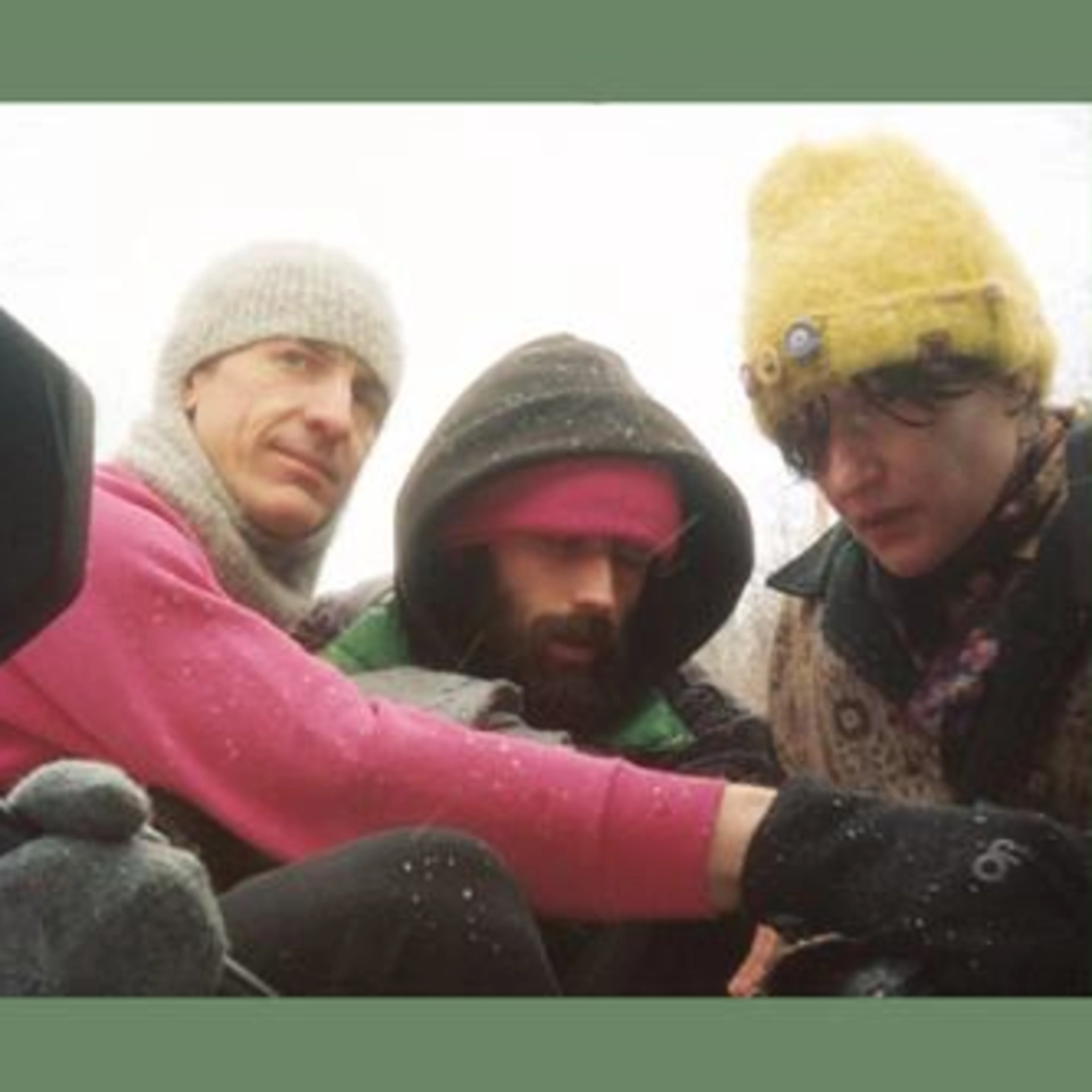 Three people in winter clothing huddled together outdoors, wearing knit hats and hooded jackets in cold weather.