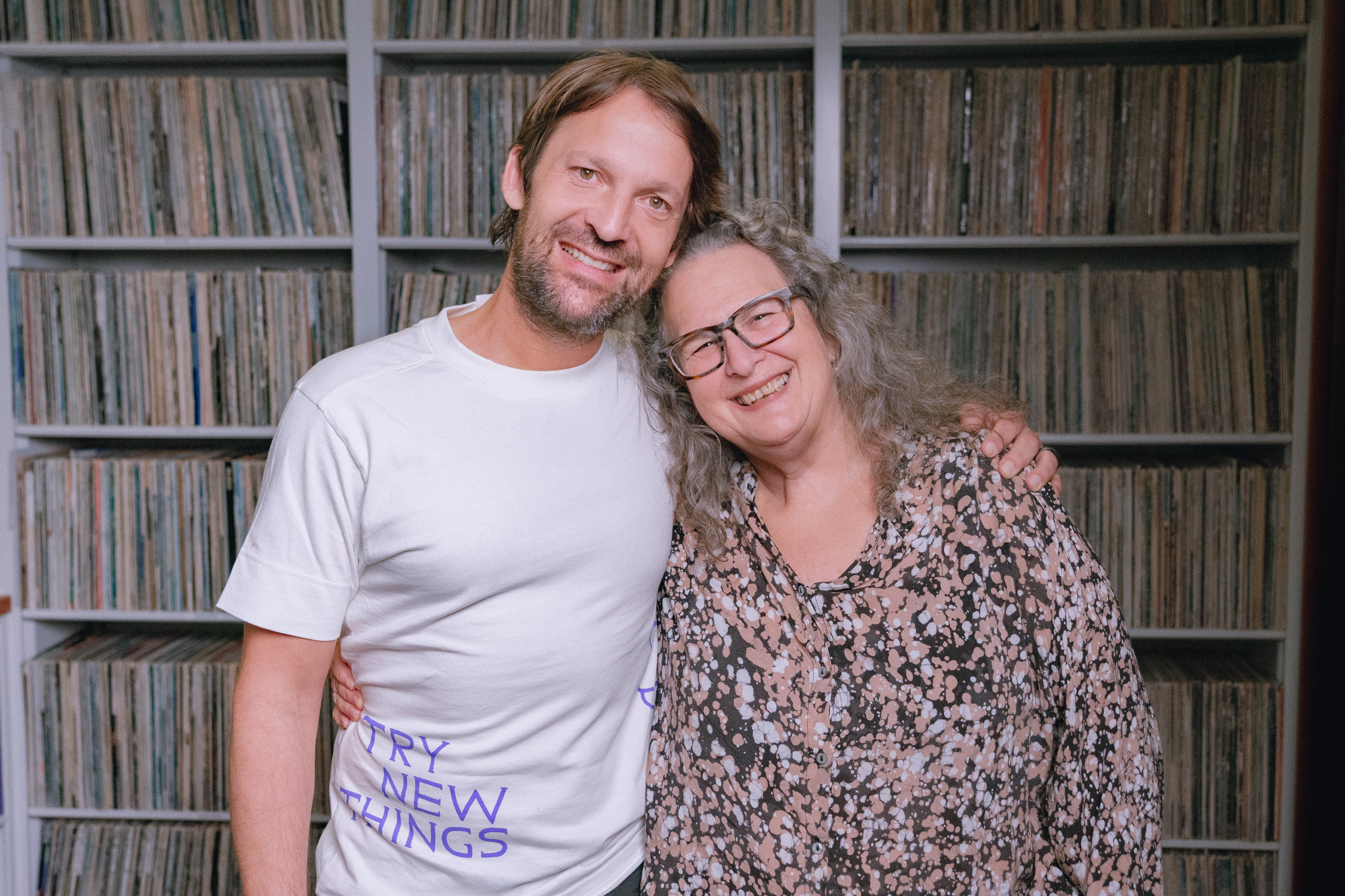 Two smiling people embracing in front of extensive vinyl record shelves, one wearing a "Try New Things" t-shirt.