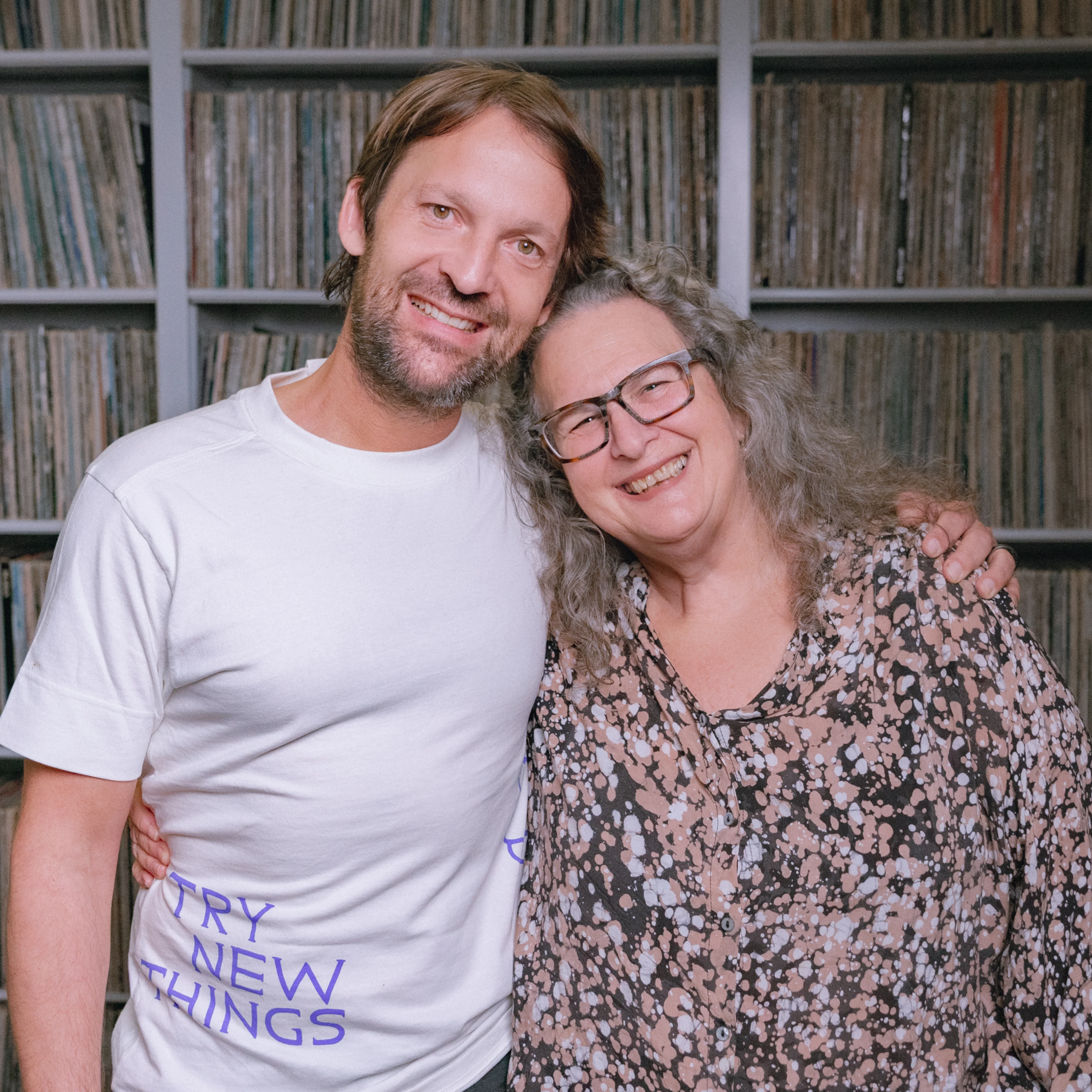 Two smiling people embracing in front of extensive vinyl record shelves, one wearing a "Try New Things" t-shirt.