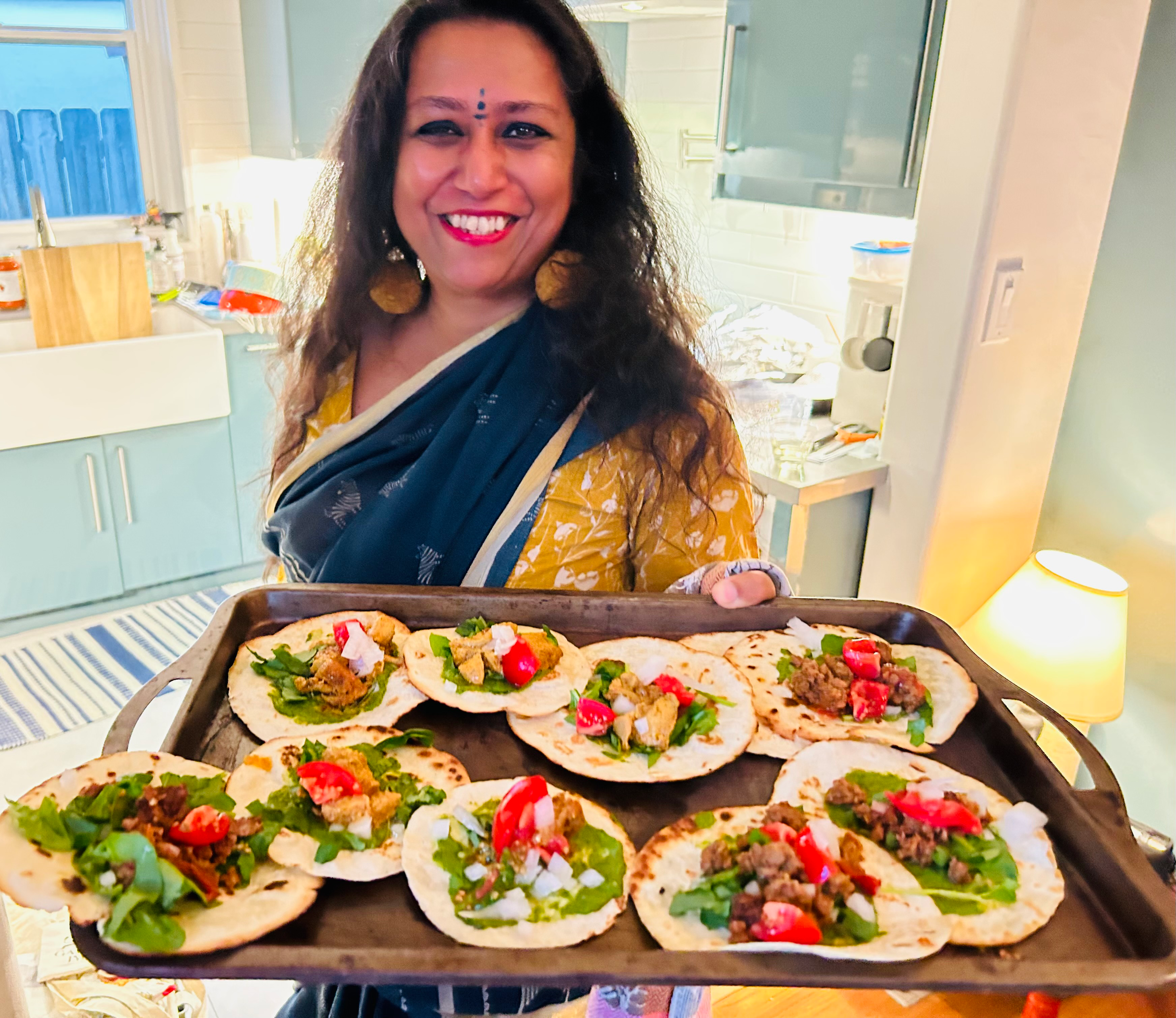 A smiling woman in a blue and yellow sari holds a tray of topped flatbreads with greens, tomatoes, and meat in a kitchen.