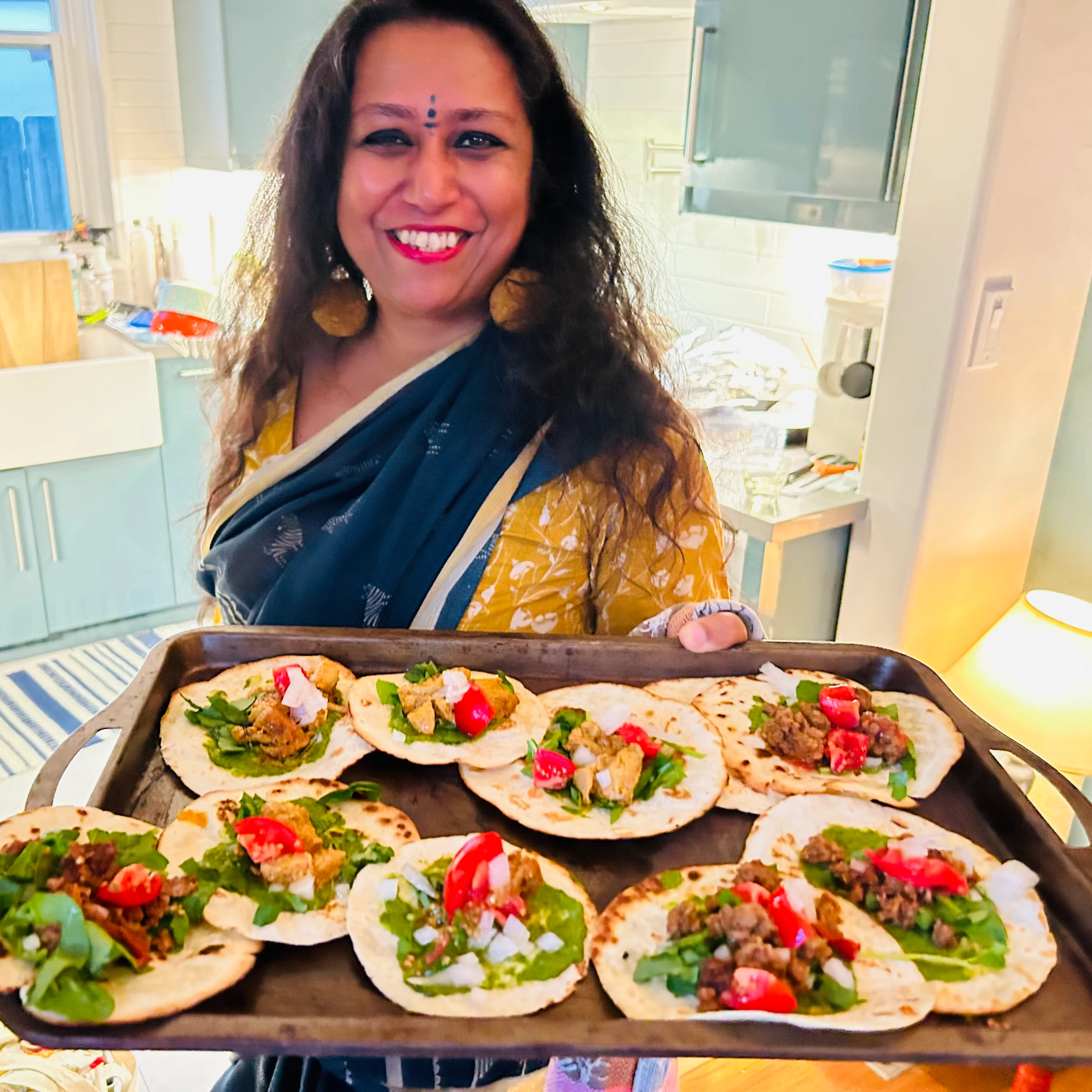 A smiling woman in a blue and yellow sari holds a tray of topped flatbreads with greens, tomatoes, and meat in a kitchen.