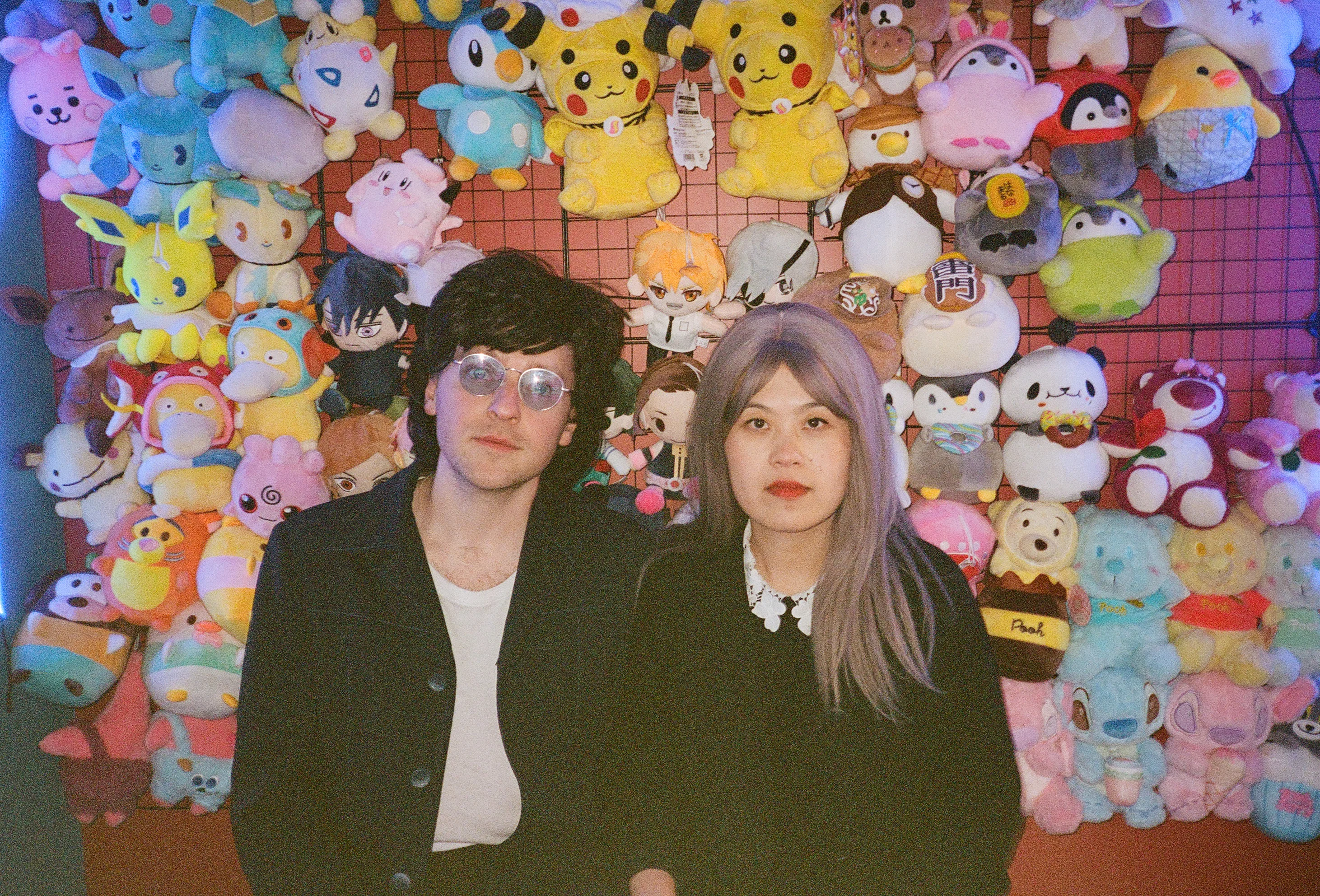 The band Chinese American Bear pose in front of a colorful wall of plush toys, including Pikachu and other anime characters.