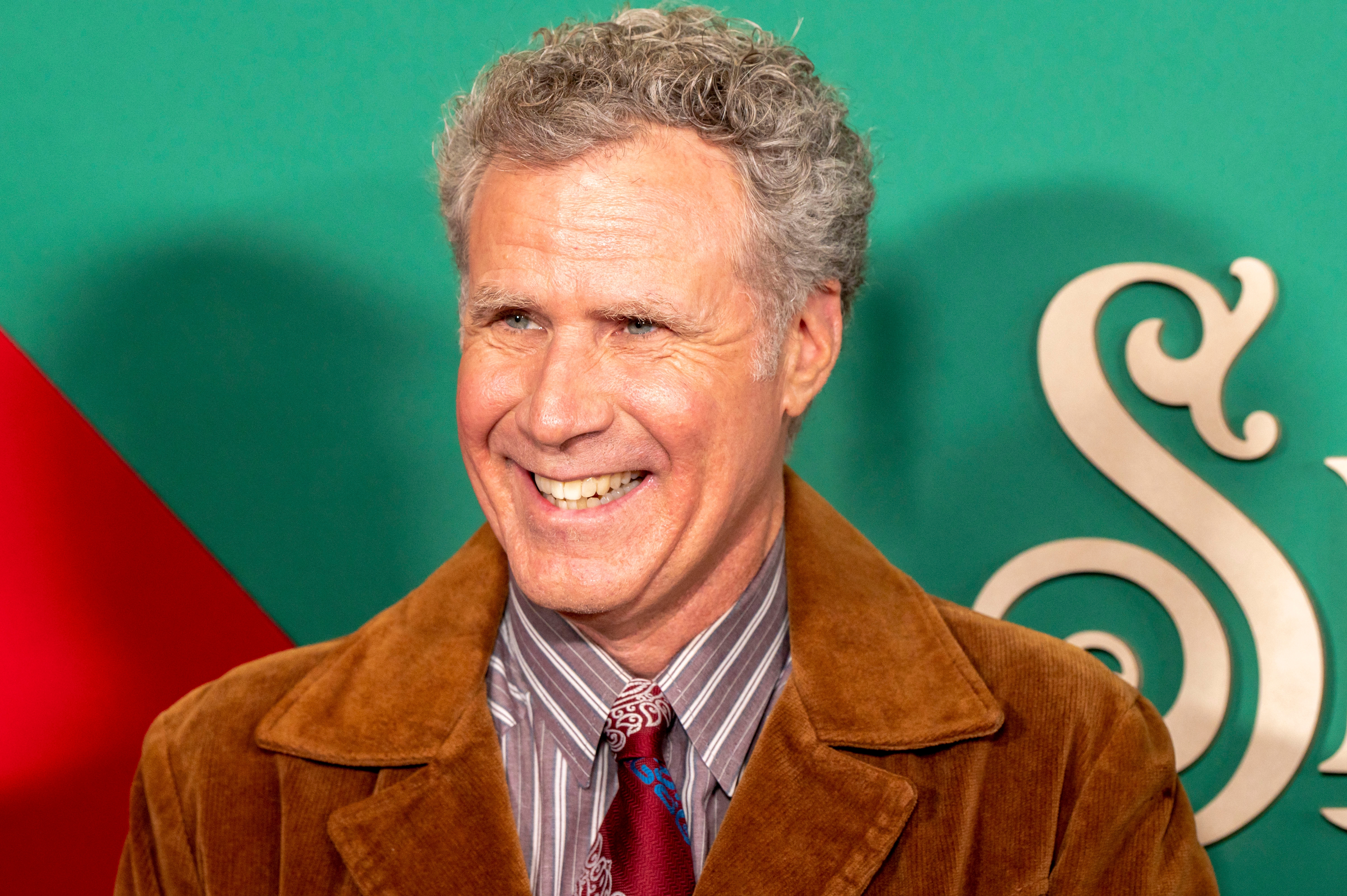 Person with gray curly hair smiling in brown jacket against green background with decorative gold element visible.