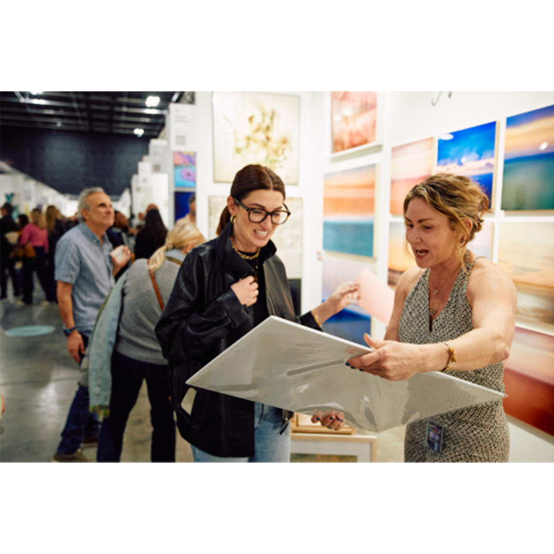 Two women discussing artwork at a gallery exhibition, with colorful framed pieces displayed on white walls behind them.