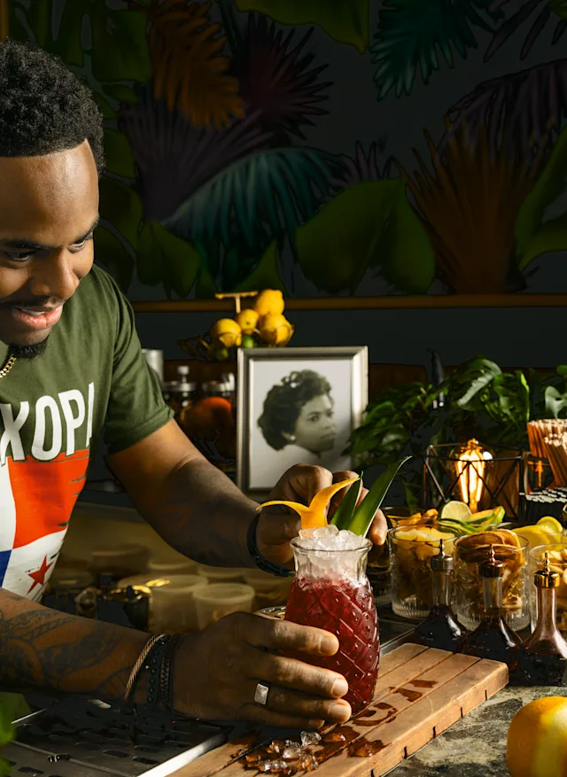 A smiling man garnishes a tropical red cocktail on a wooden bar surrounded by fruits and candles with tropical wallpaper behind him.