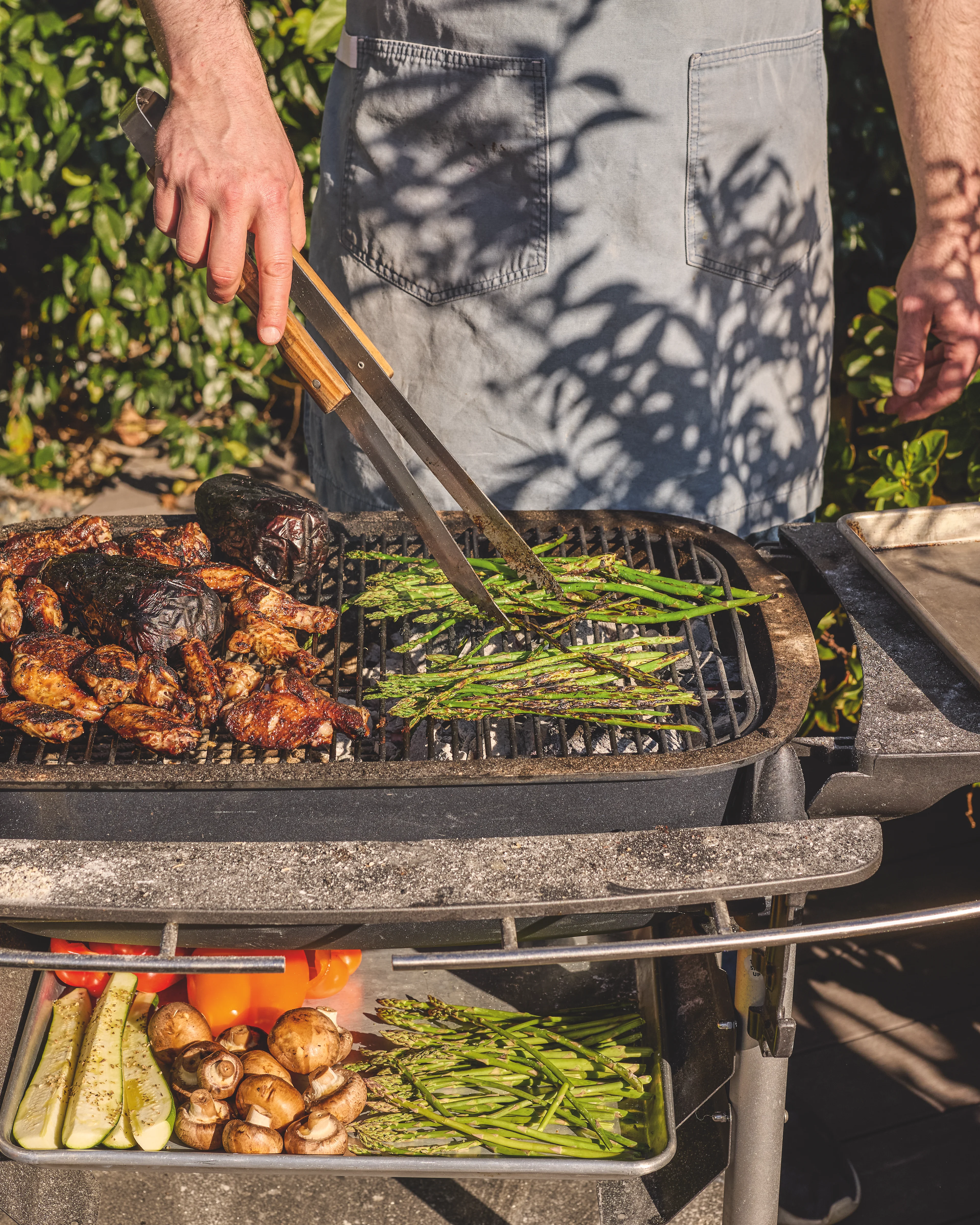 A person in a gray apron grills asparagus and chicken on an outdoor grill, with fresh vegetables stored on the shelf below.