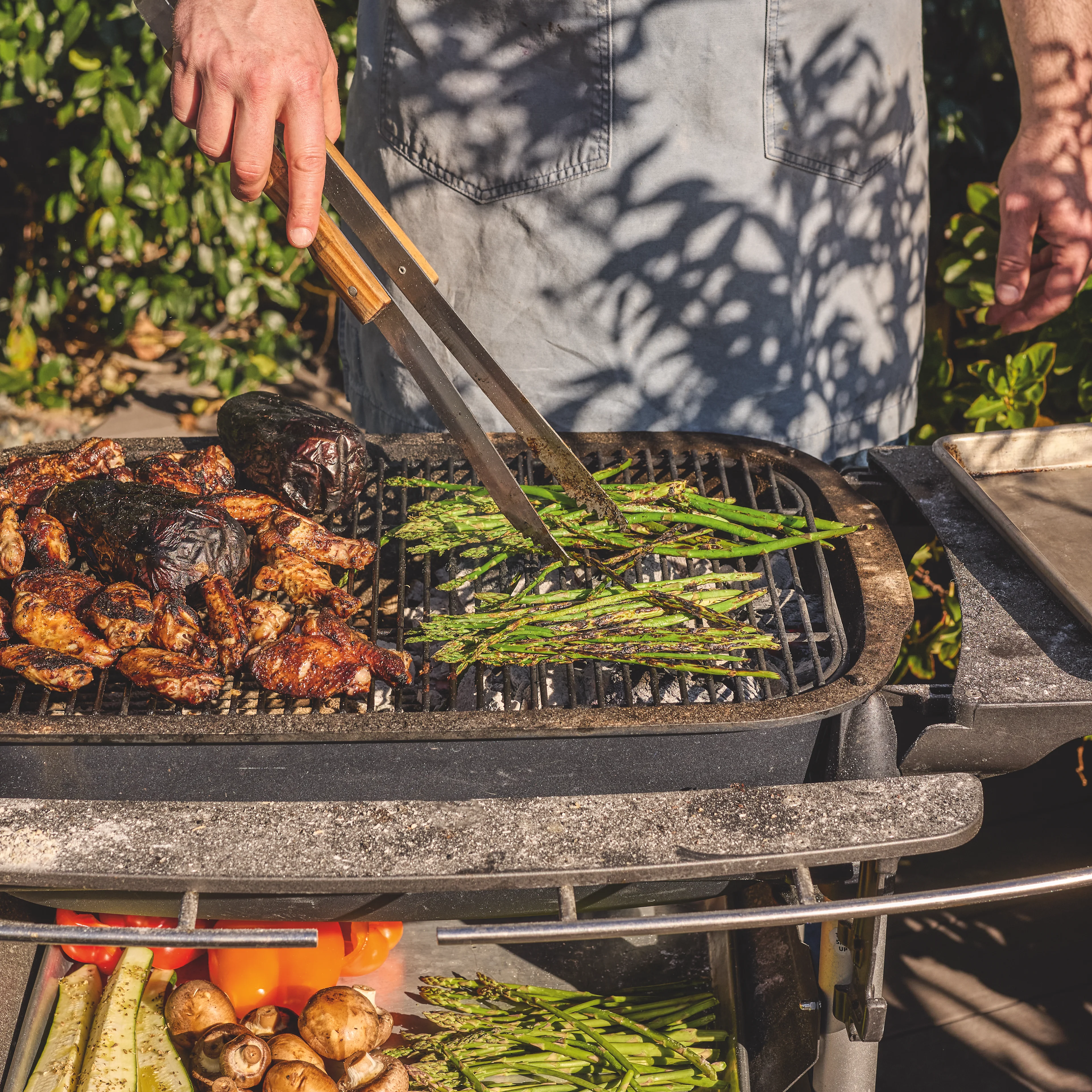 A person in a gray apron grills asparagus and chicken on an outdoor grill, with fresh vegetables stored on the shelf below.