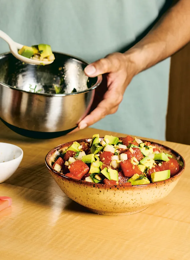 Person preparing fresh salad with avocado and tomato chunks in ceramic bowl while adding ingredients from metal mixing bowl.