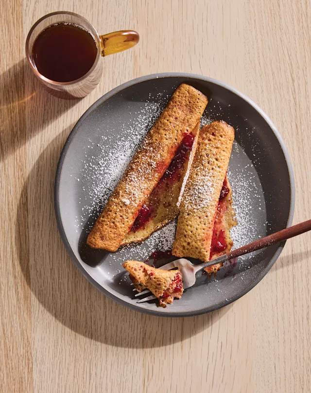 Rolled crepes filled with red berry jam, dusted with powdered sugar on a gray plate, with a fork and cup of tea nearby.