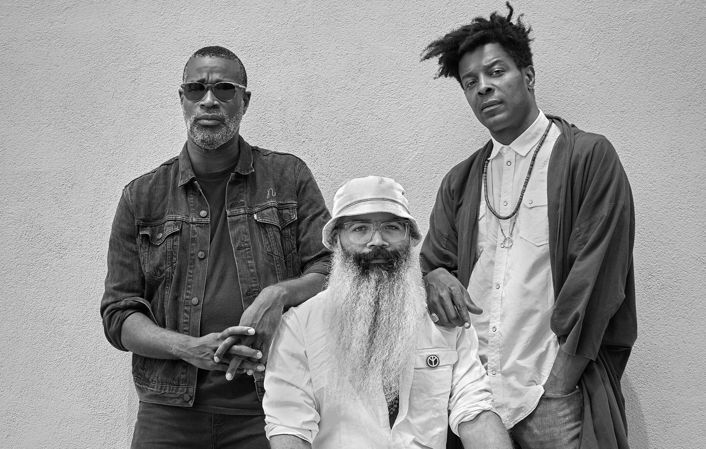 Three men posing in black and white portrait - one with sunglasses, one with bucket hat and beard, one with curly hair.