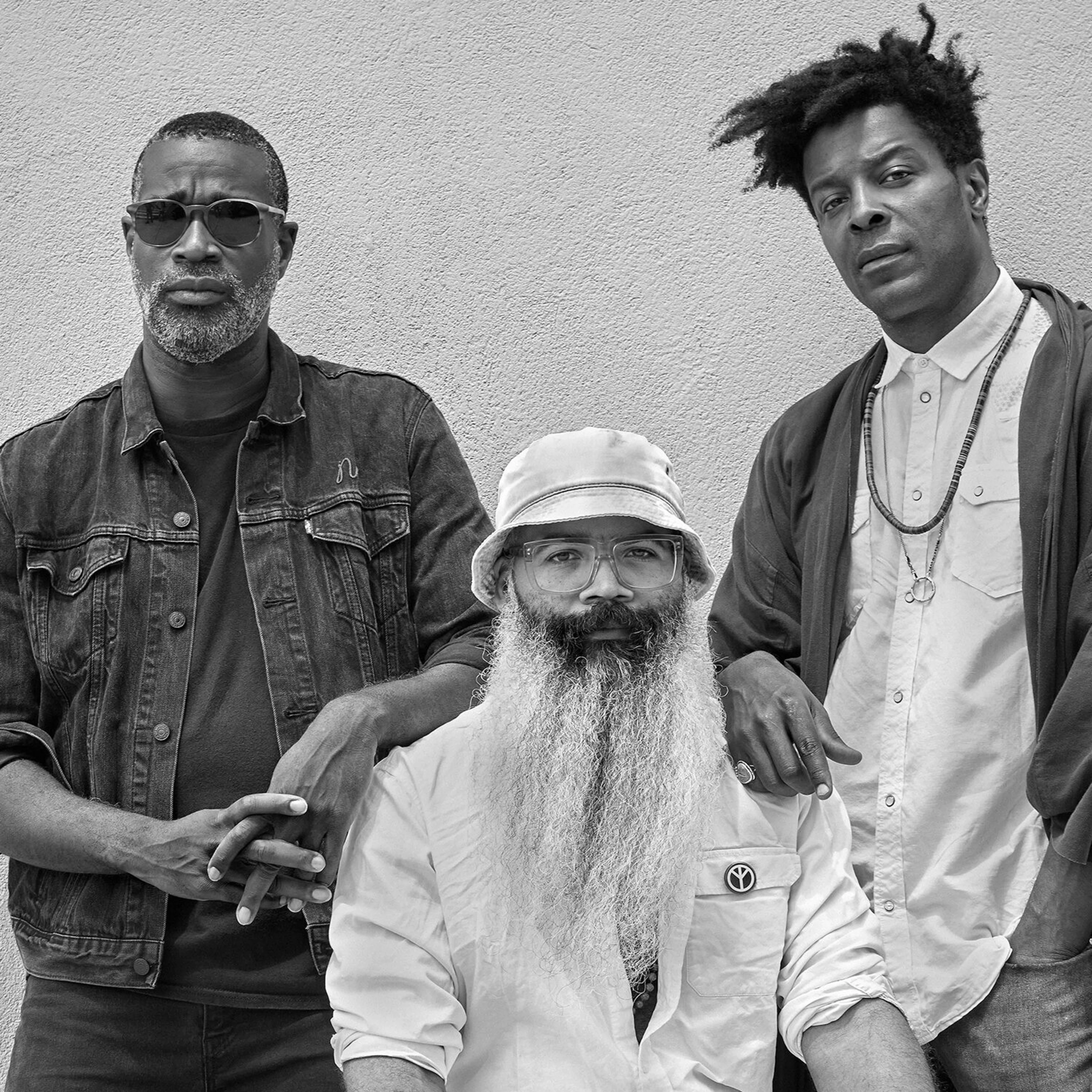 Three men posing in black and white portrait - one with sunglasses, one with bucket hat and beard, one with curly hair.