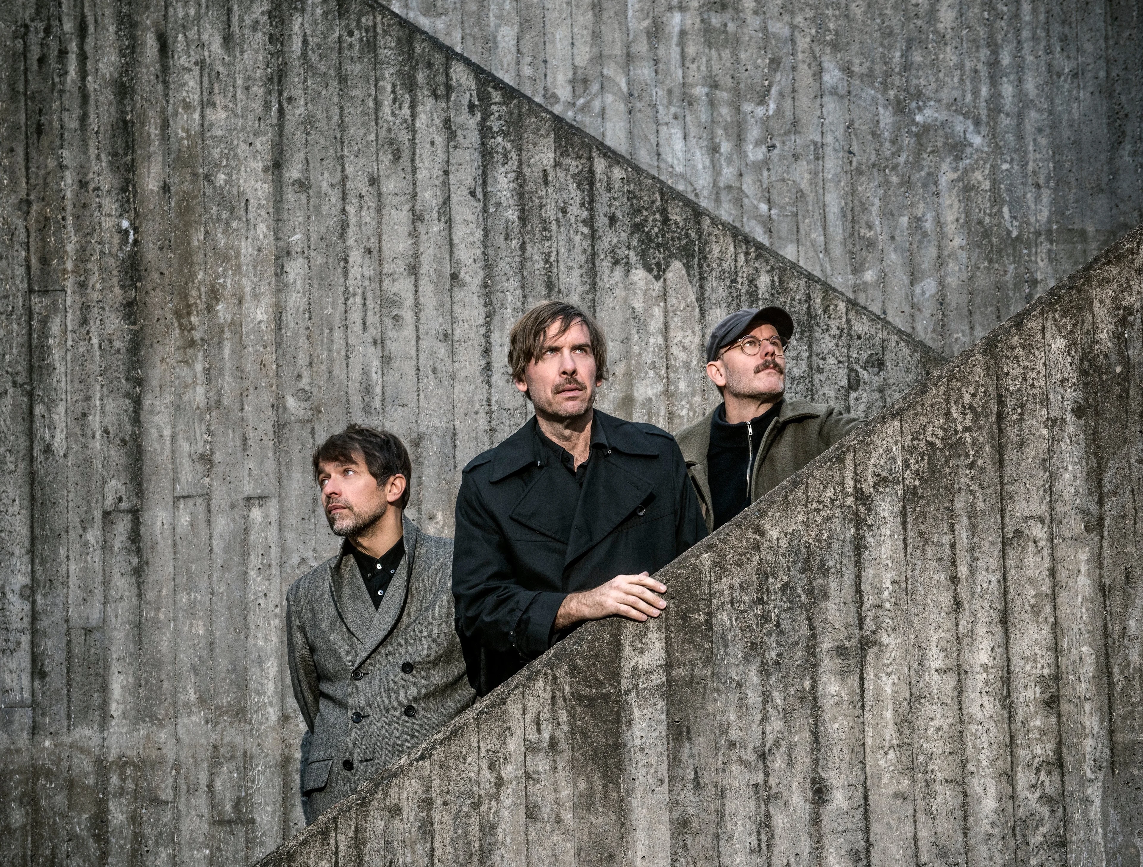 Three men in dark clothing stand by a concrete staircase, gazing upward against a gray brutalist wall.
