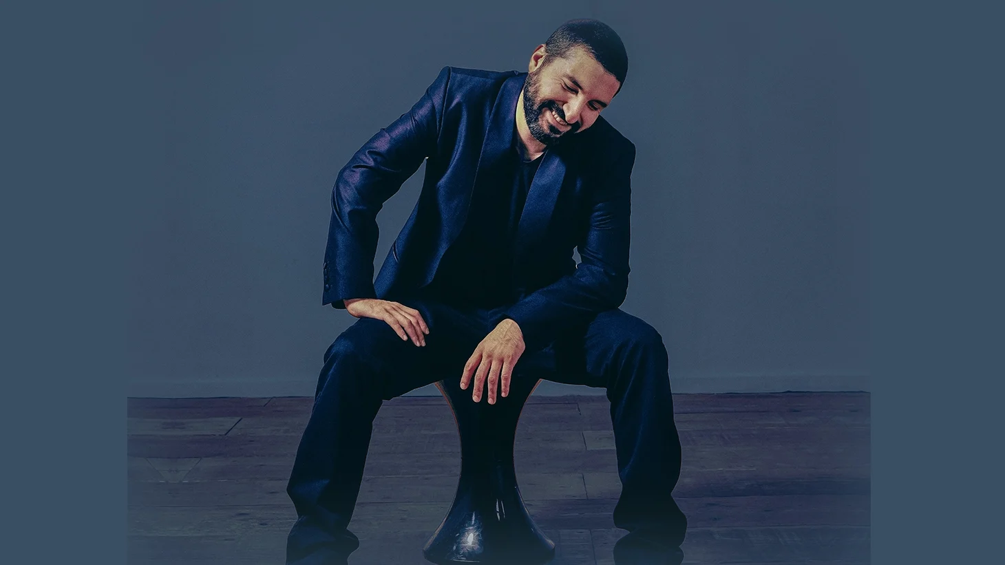 A smiling Ibrahim Maalouf in a navy blue suit sits on a modern stool, leaning forward against a blue-gray background.
