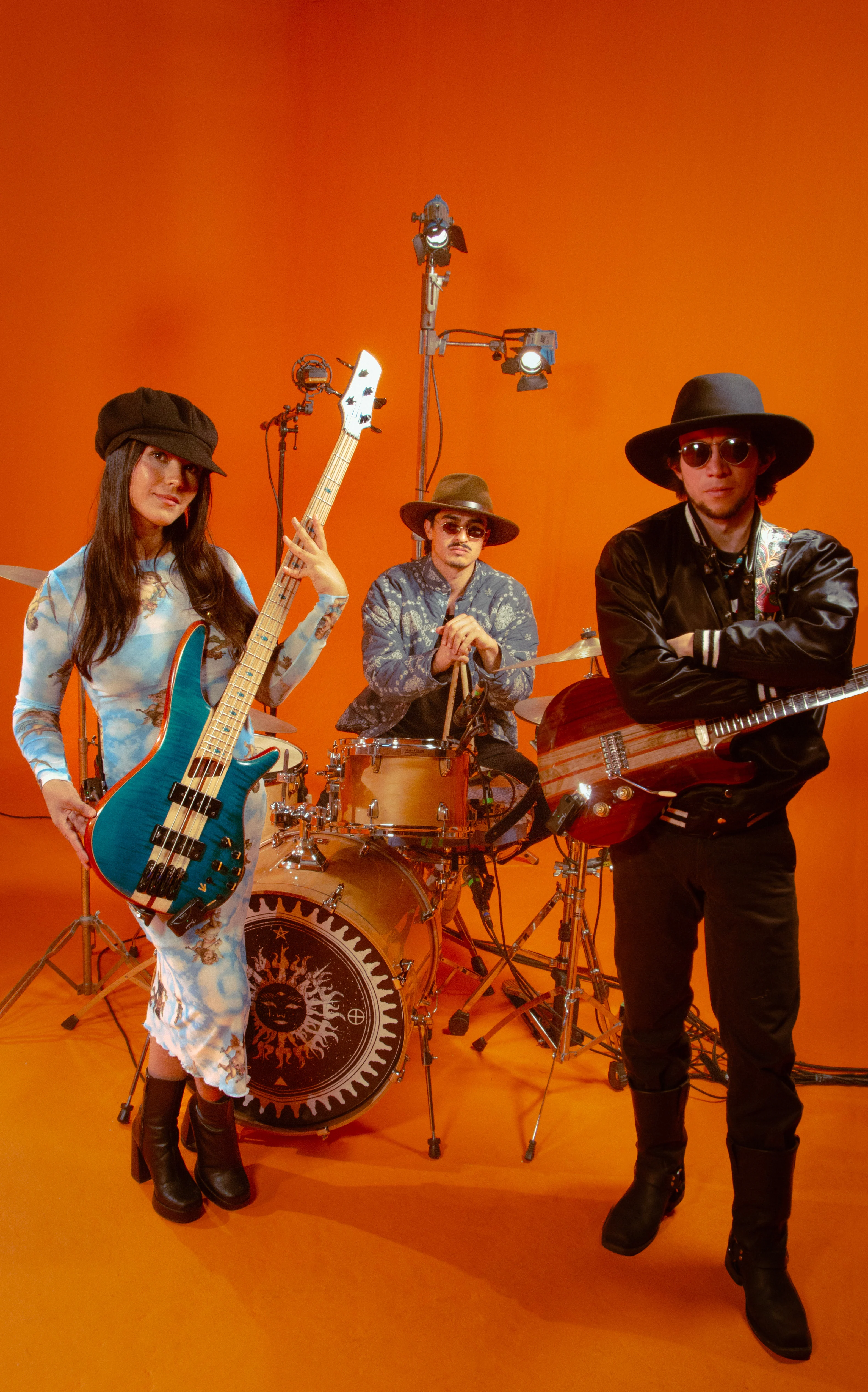 Three-piece band poses against an orange backdrop; woman holds blue bass guitar, drummer sits behind kit, guitarist stands with red guitar.