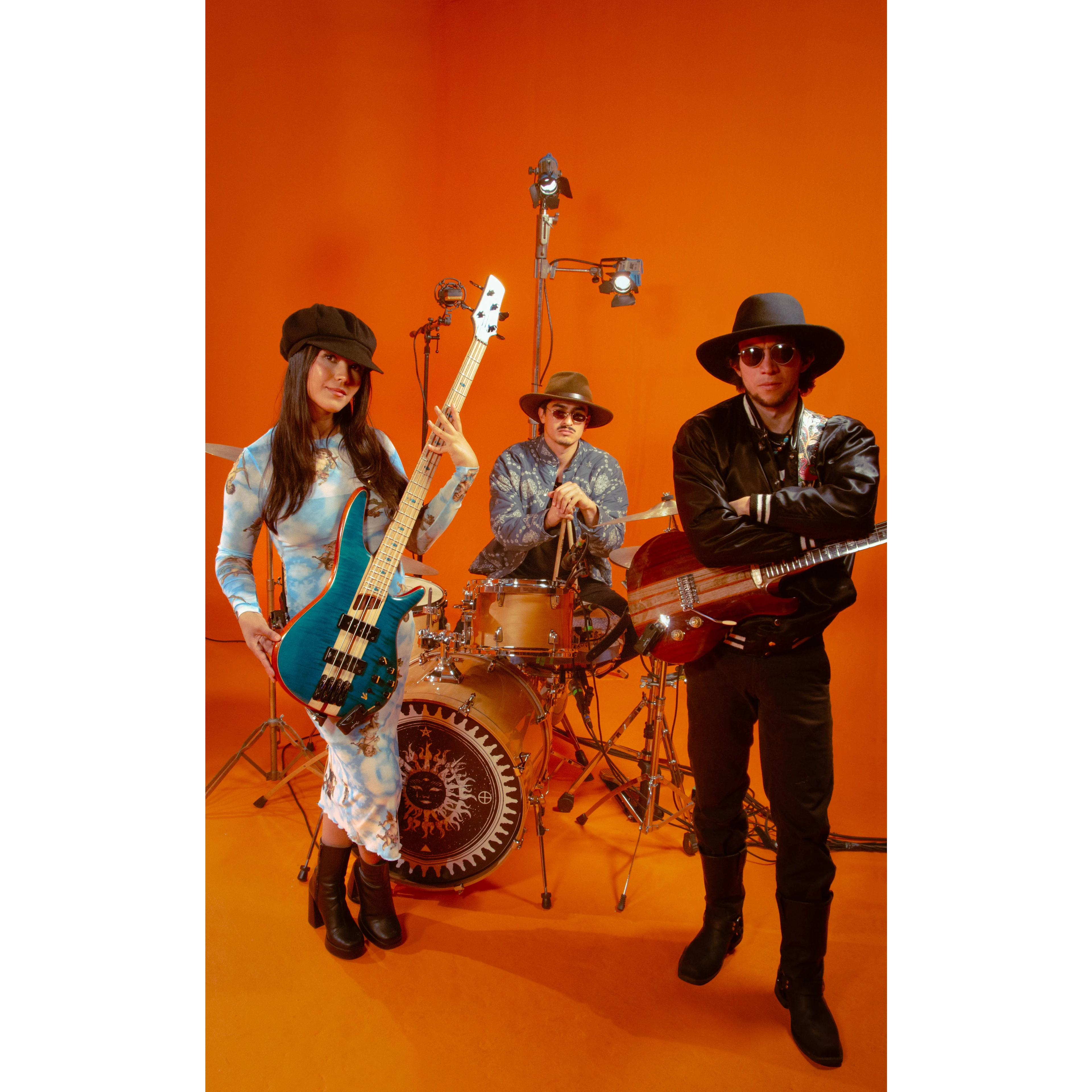 Three-piece band poses against an orange backdrop; woman holds blue bass guitar, drummer sits behind kit, guitarist stands with red guitar.