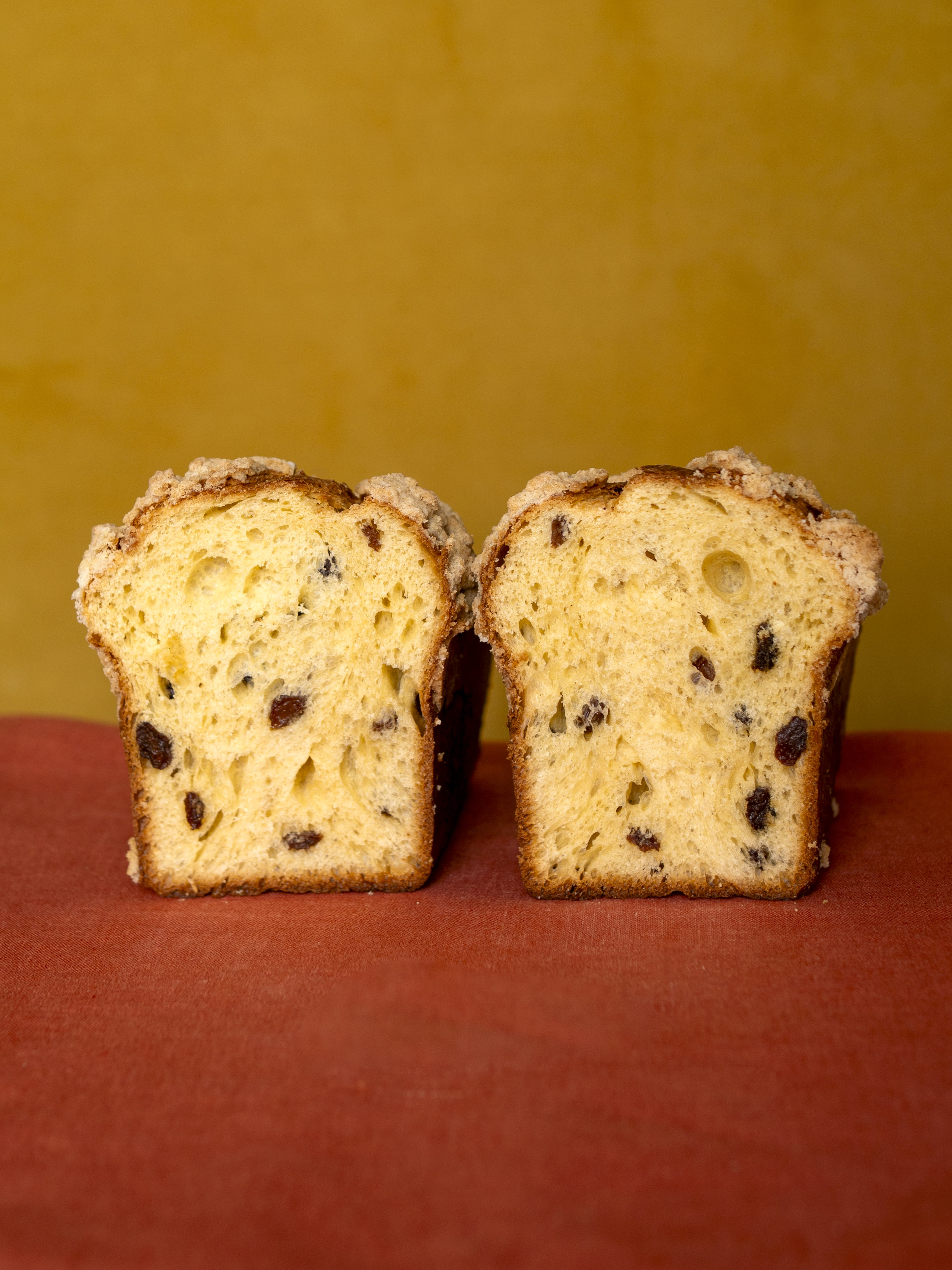 Two slices of golden panettone with raisins displayed on a red surface against a mustard yellow background.