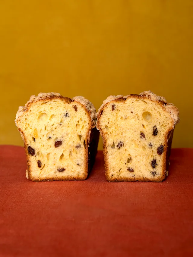 Two slices of golden panettone with raisins displayed on a red surface against a mustard yellow background.