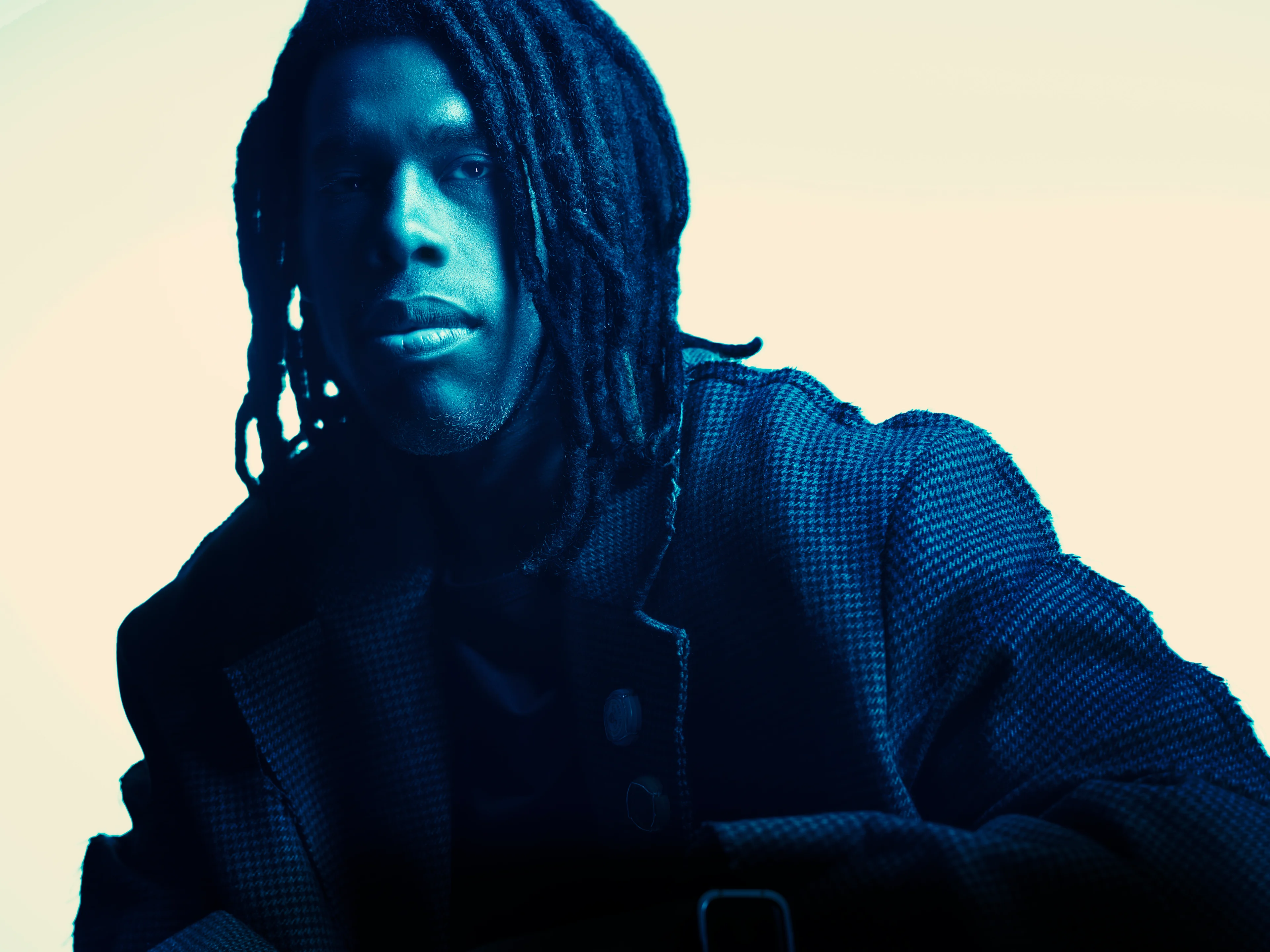 A man with long dreadlocks wearing a houndstooth jacket, photographed with a cool blue tint against a light background.