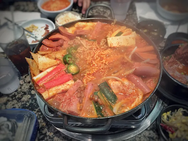 Steaming pot of Korean budae jjigae (army stew) with ramen, spam, tofu, and vegetables in spicy red broth on a tabletop burner.