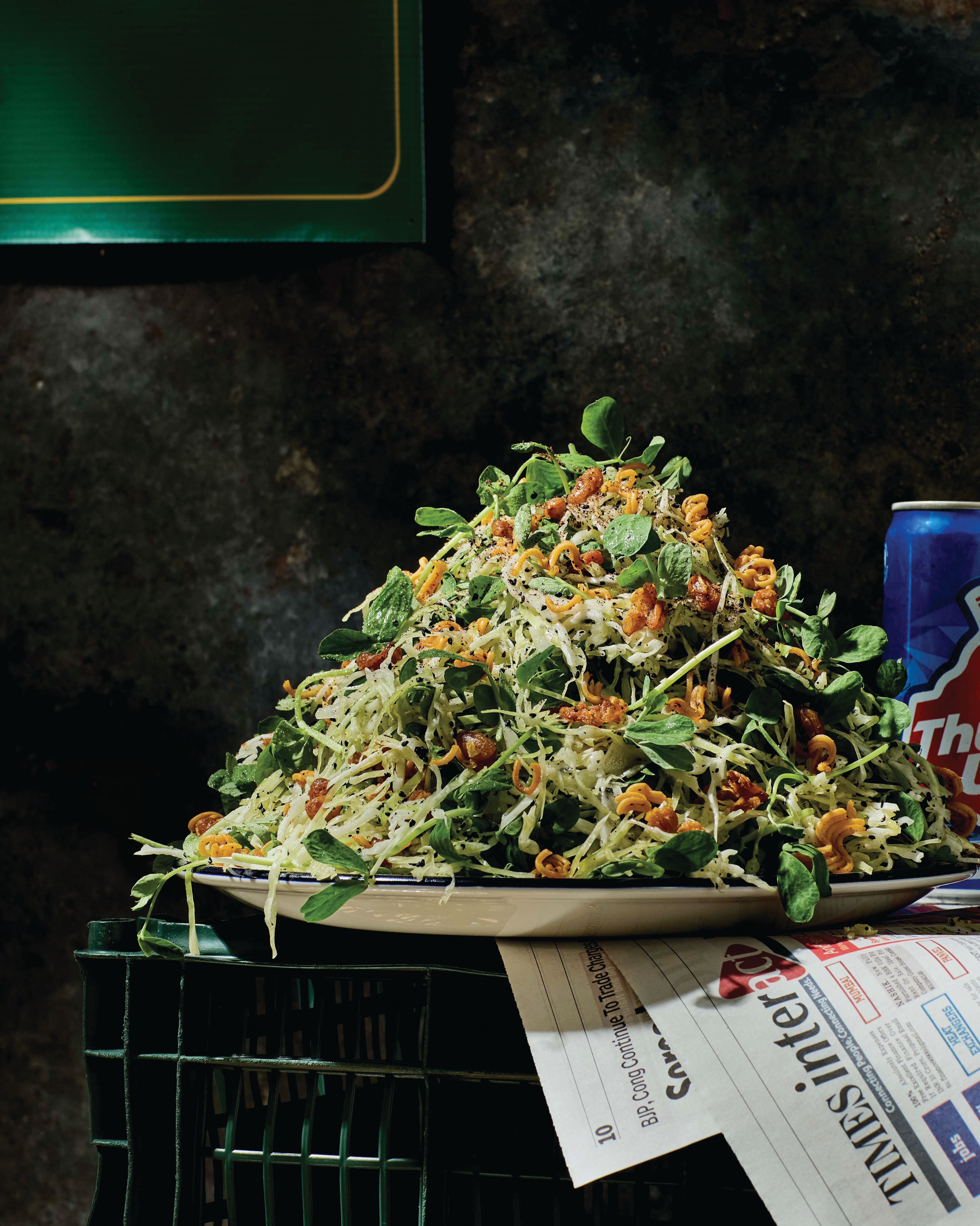A large mound of fresh coleslaw salad with greens and crunchy toppings on a white plate, next to a soda can and newspaper.