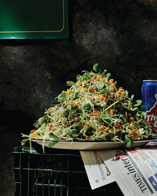 A large mound of fresh coleslaw salad with greens and crunchy toppings on a white plate, next to a soda can and newspaper.
