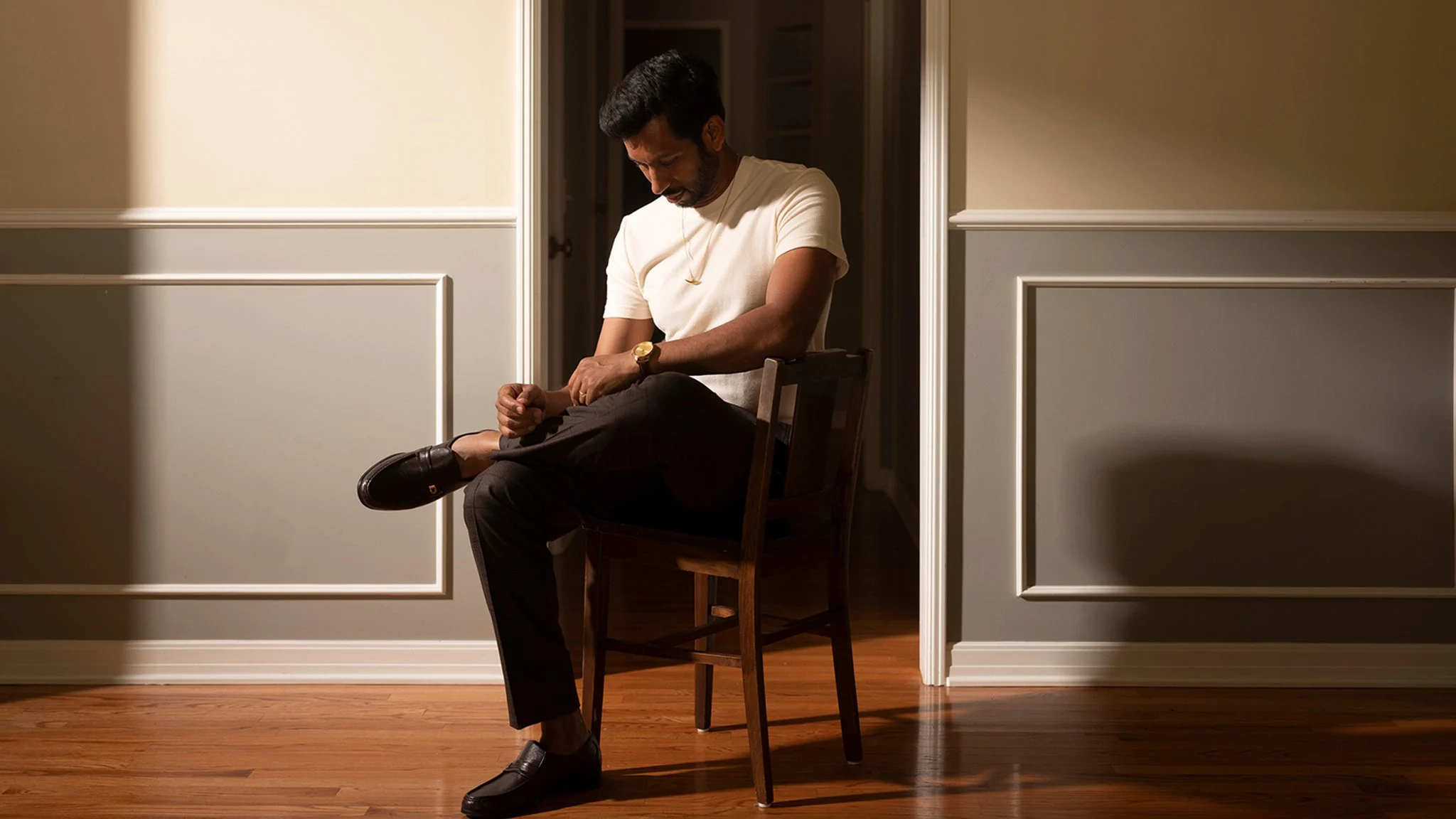 Hrishikesh Hirway in white t-shirt sitting on wooden chair in doorway, adjusting dark shoes, warm lighting on wooden floor.