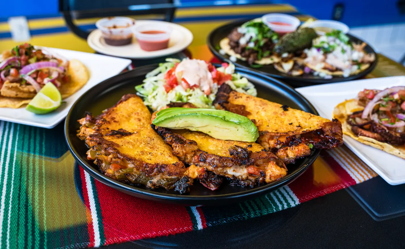 Colorful Mexican food spread with crispy quesadillas topped with avocado, tacos, and small dishes of salsa on a striped tablecloth.