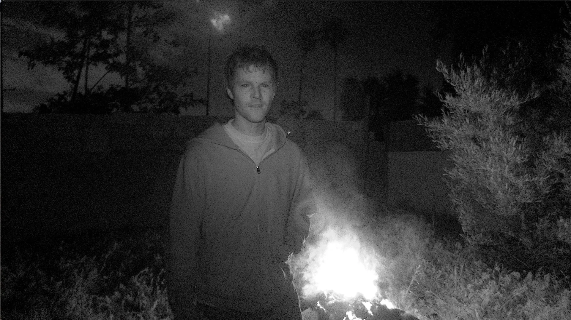 A young man in a hoodie stands outdoors at night near a small fire, with trees and a building visible in the background.